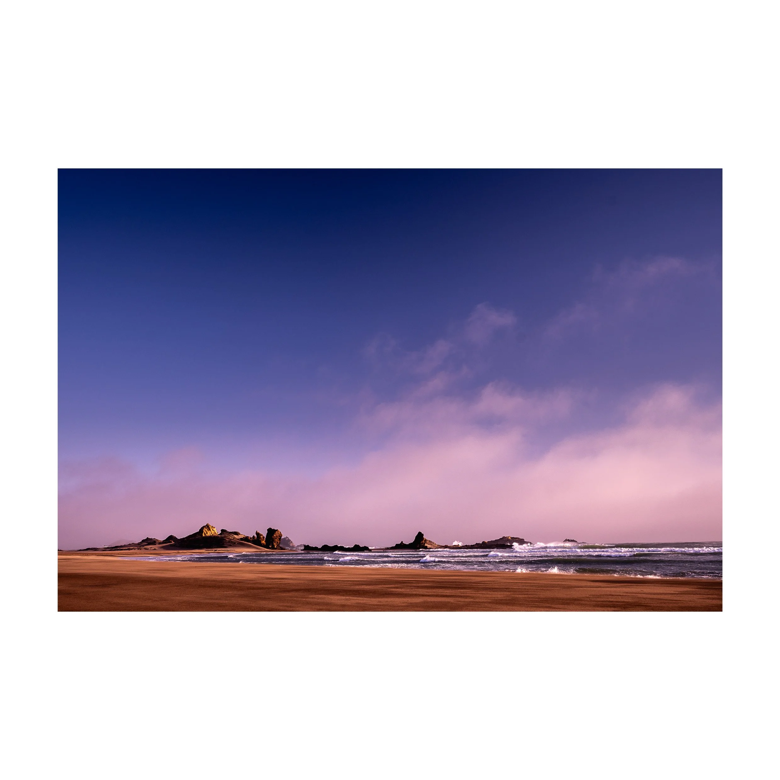 A beach with rocks and waves under a purple and blue sky.