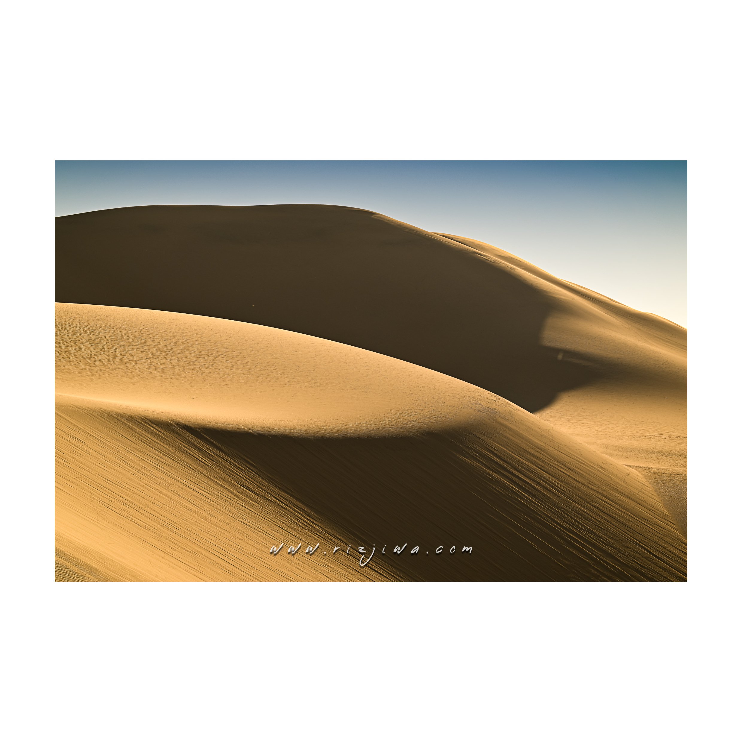 Sand dunes in a desert with smooth, curved shapes, and a clear blue sky in the background.