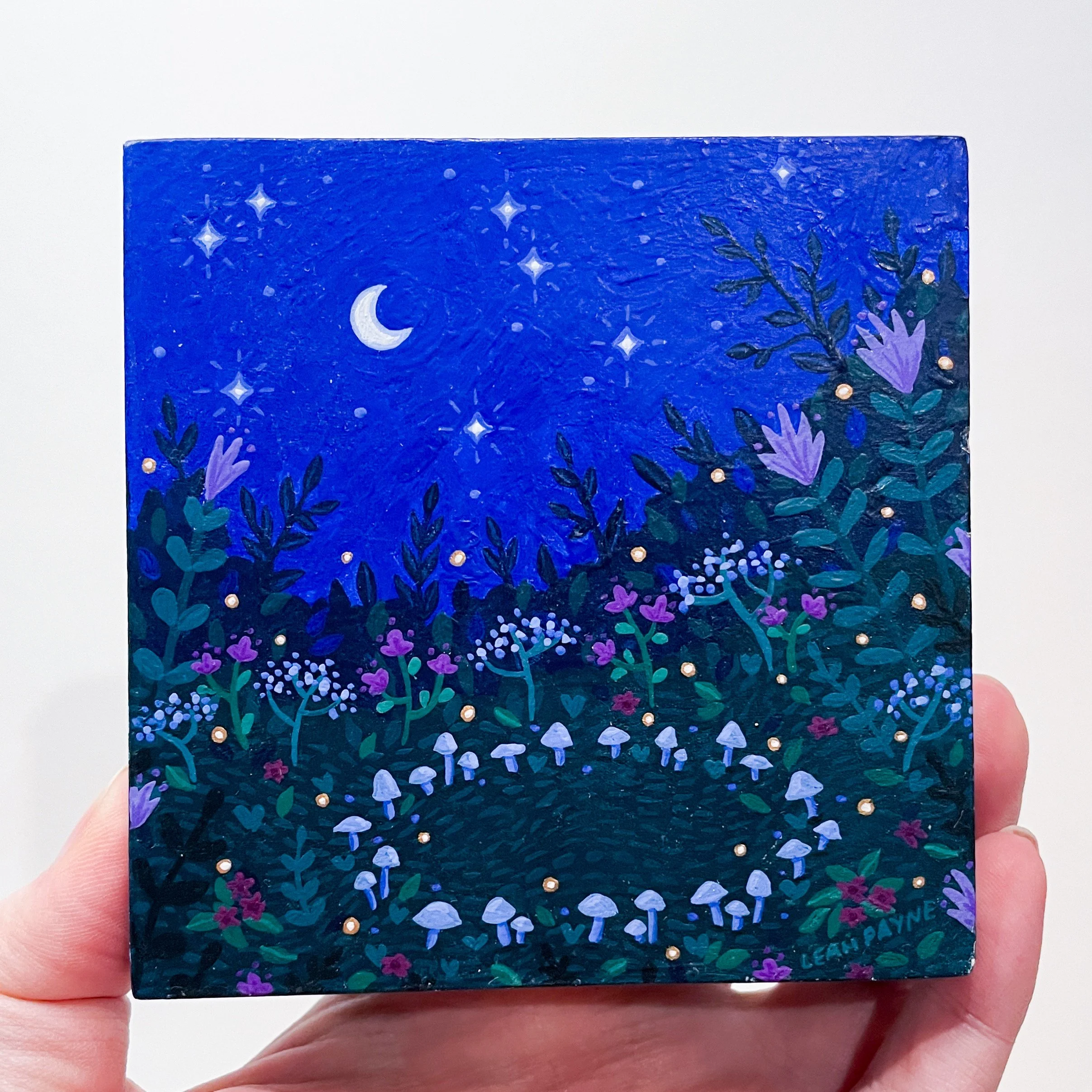 a hand holds up a 4"x4" painting of a ring of mushrooms in a flower field under a midnight blue night sky