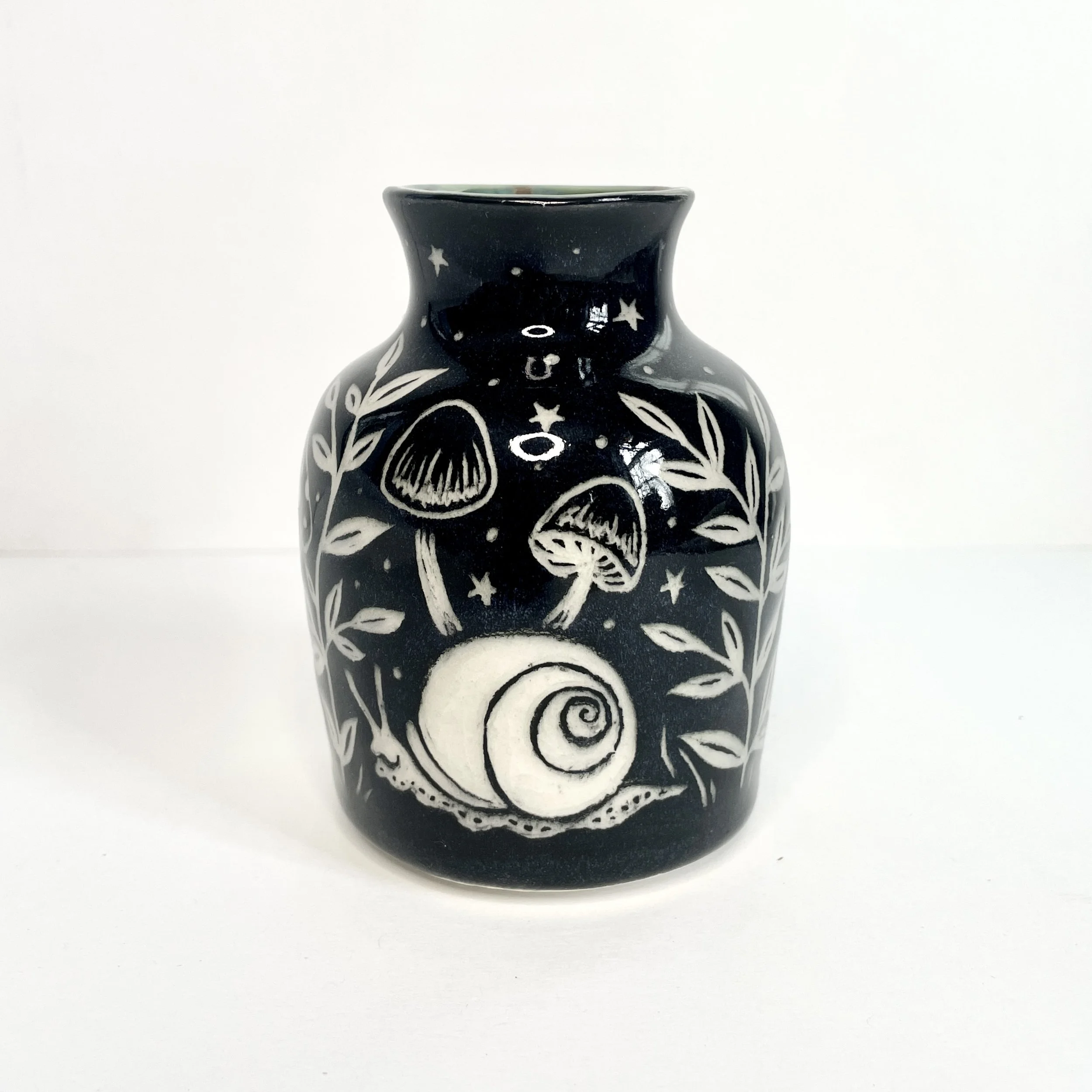 a ceramic bud vase. the exterior is black and has white snails, mushrooms and leaves. the interior has green around the rim.