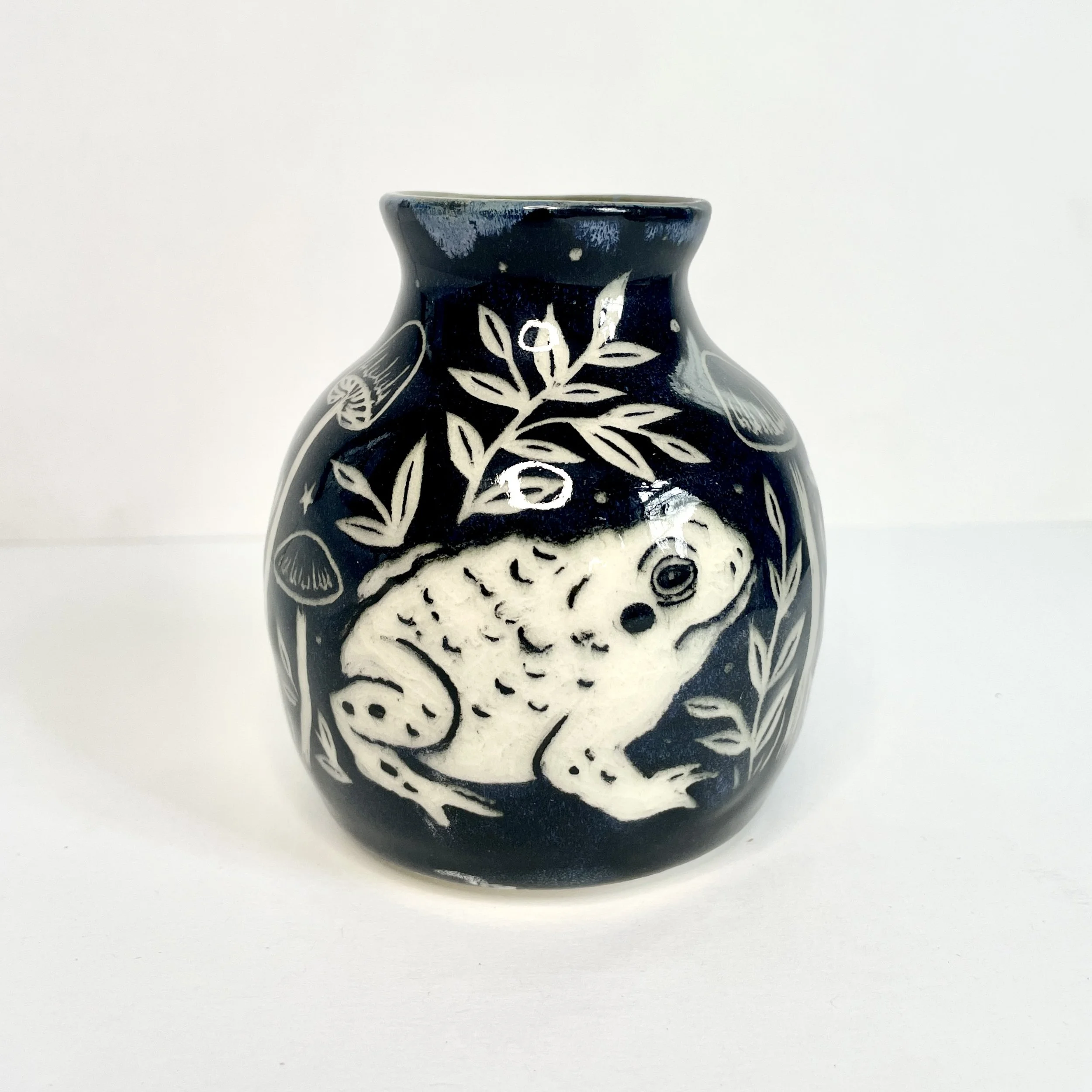 a small ceramic bud vase. the exterior is black and has white toads and leaves. the interior has green around the rim.
