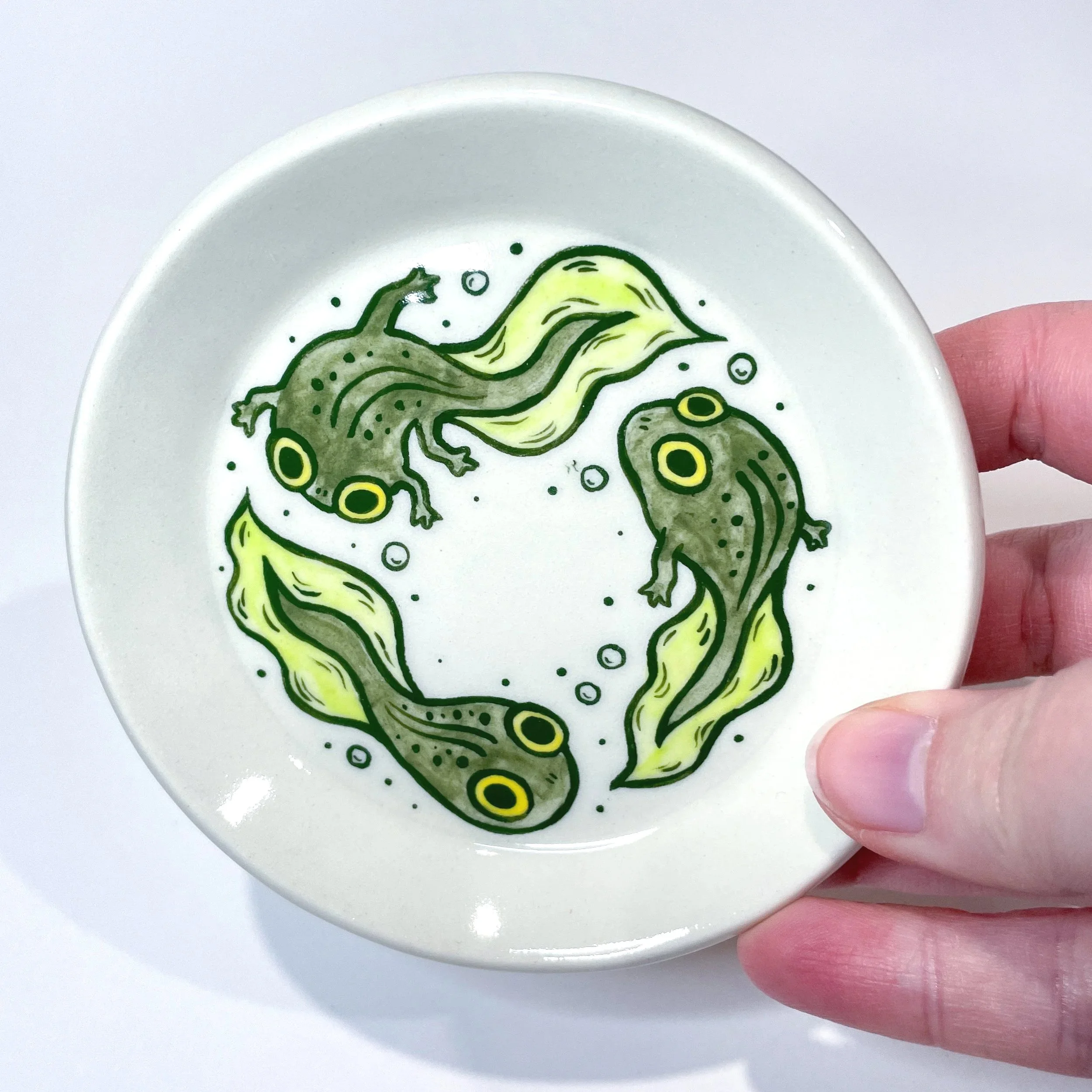 a hand holds up a small trinket dish with three tadpoles illustrated on it.