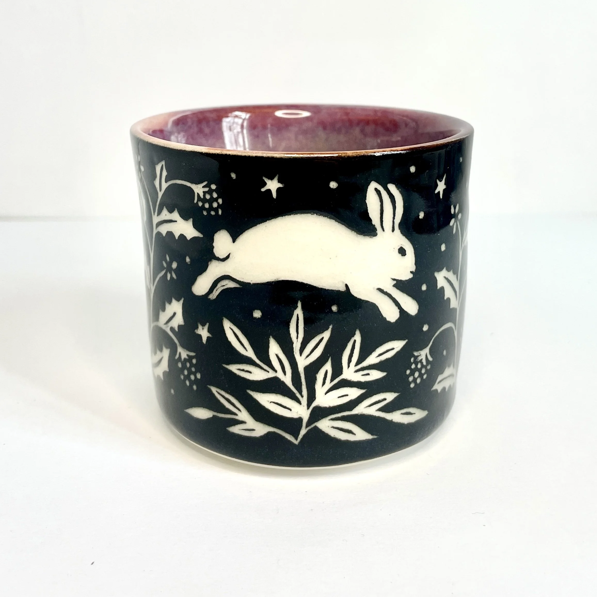 a ceramic cup. The exterior is black and has white bunnies, stars, and leaves. The interior is pink.