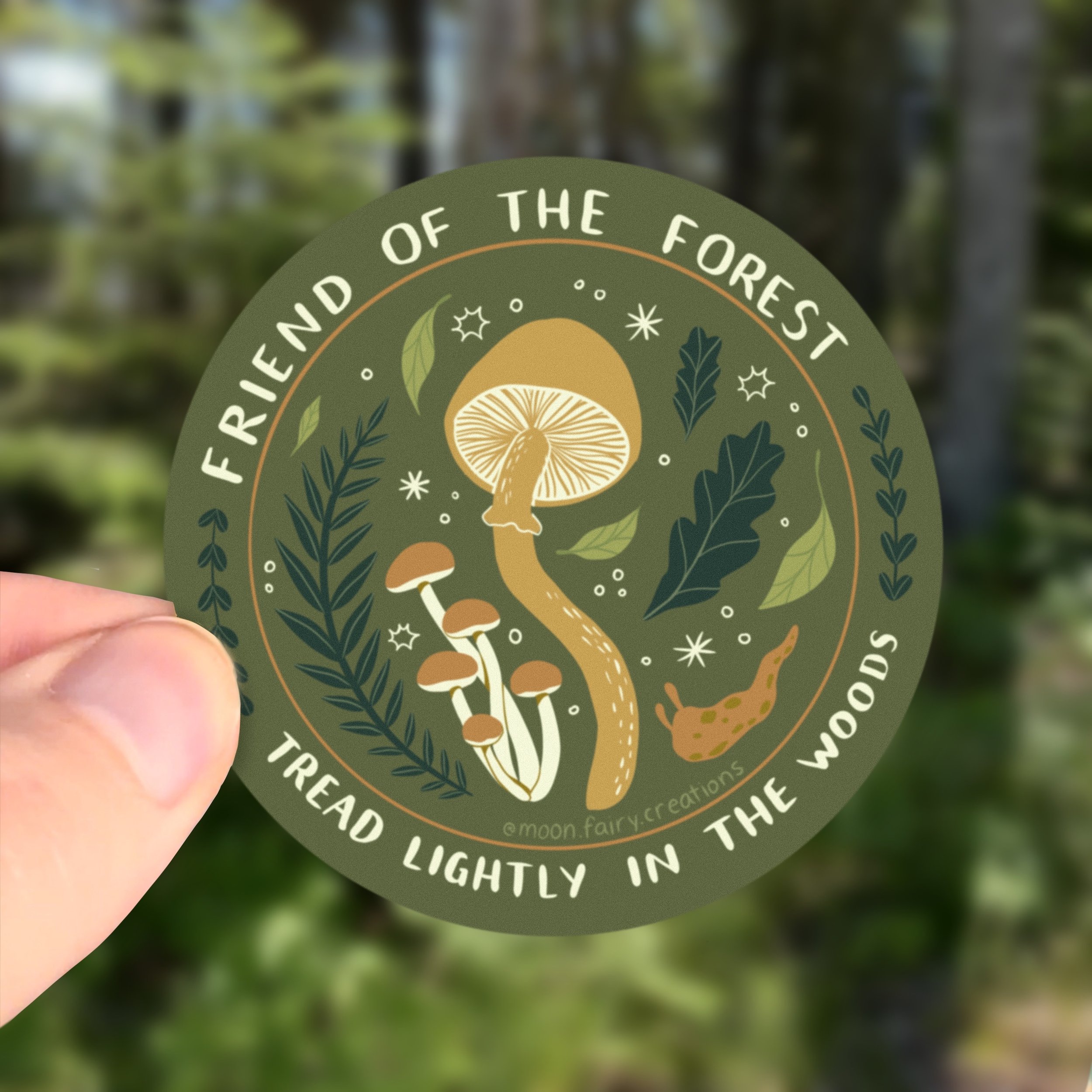 a hand holds up a sticker of forest elements and the words friend of the forest, tread lightly in the woods