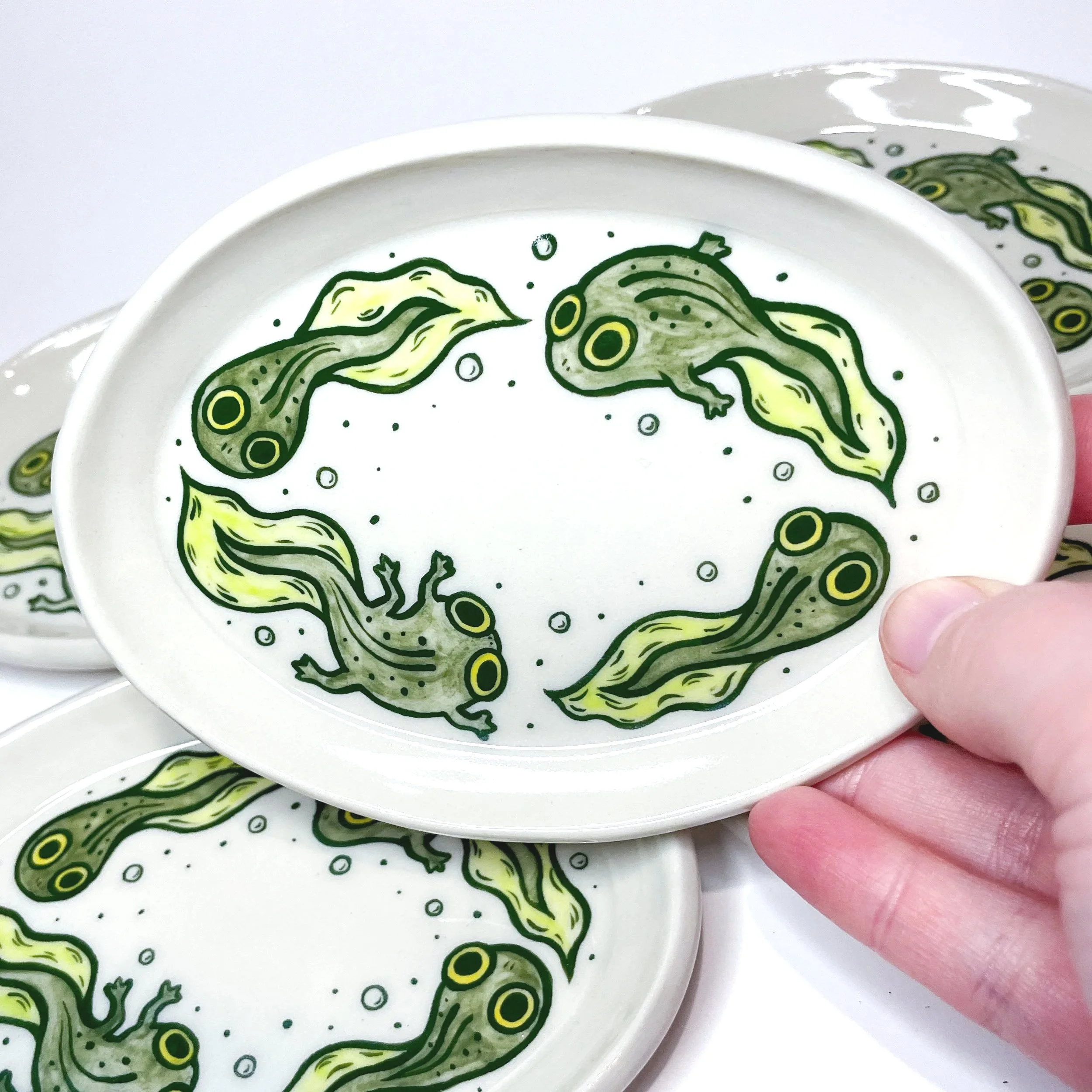 a hand holds up an oval trinket dish with four tadpoles illustrated on it. there are more of this same style trinket dish behind it.