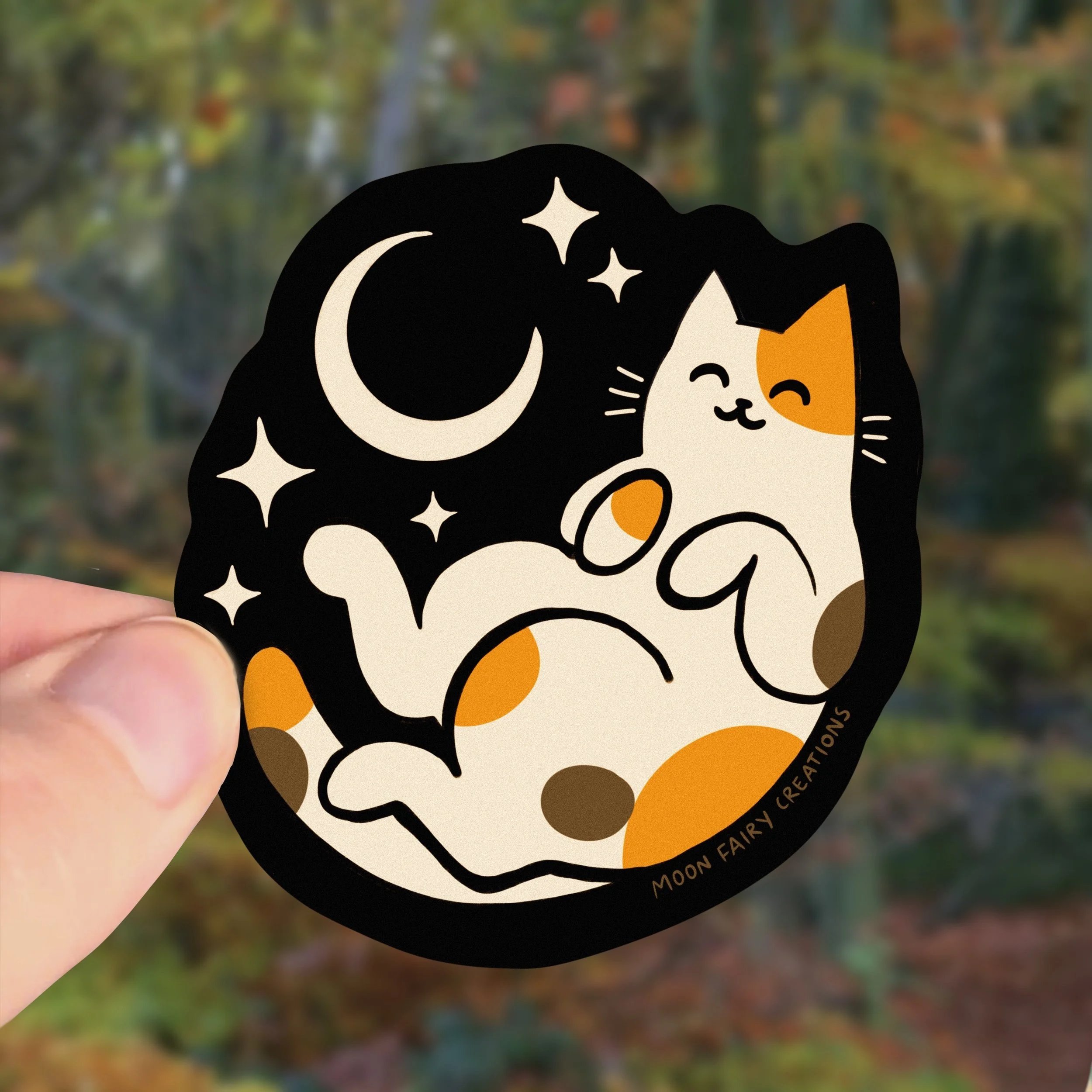 a hand holds up a sticker of a calico cat laying under the moon