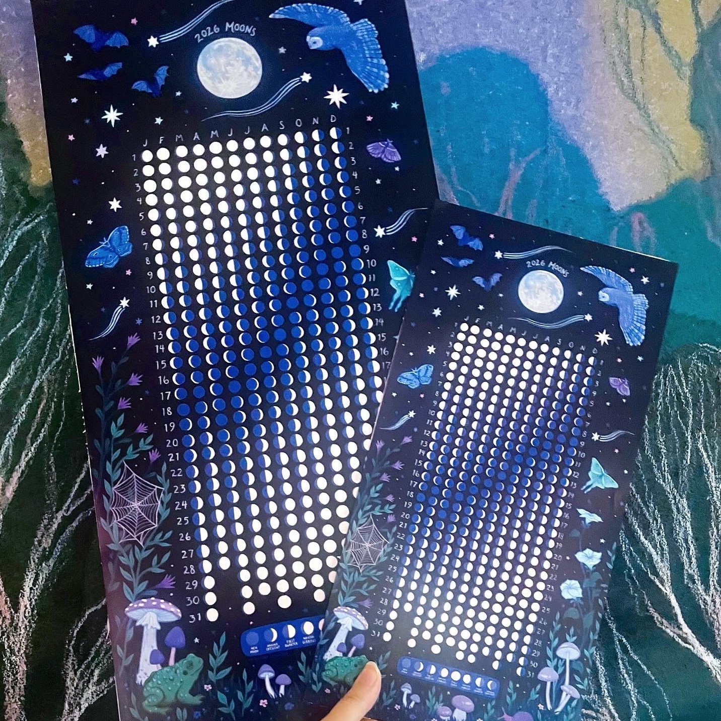 a hand holds up two different sixed moon calendars. the large one is 1 foot by two feet, the small one is 8 inches by 16 inches.