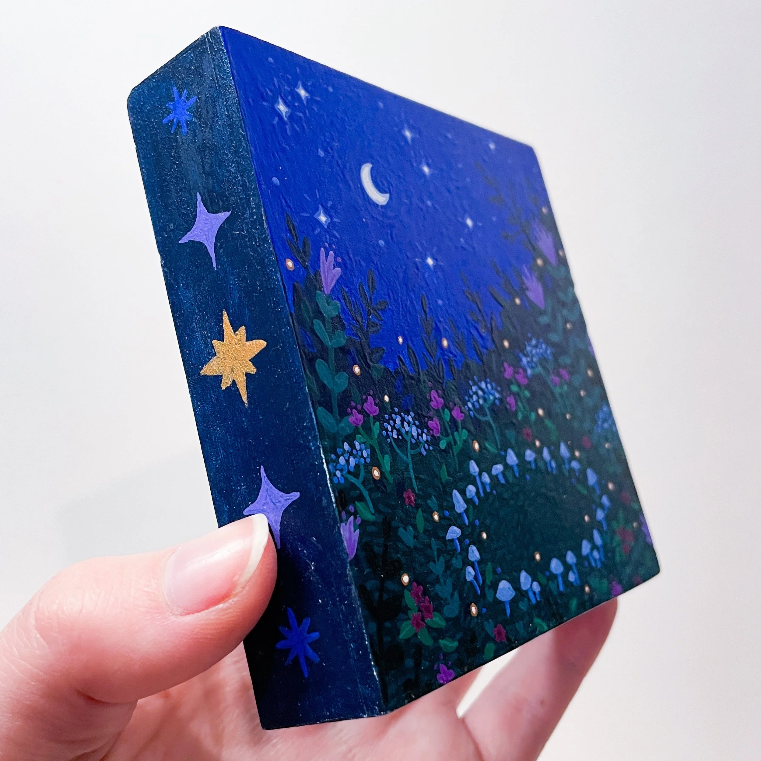 a hand holds up a 4"x4" painting of a ring of mushrooms in a flower field under a midnight blue night sky