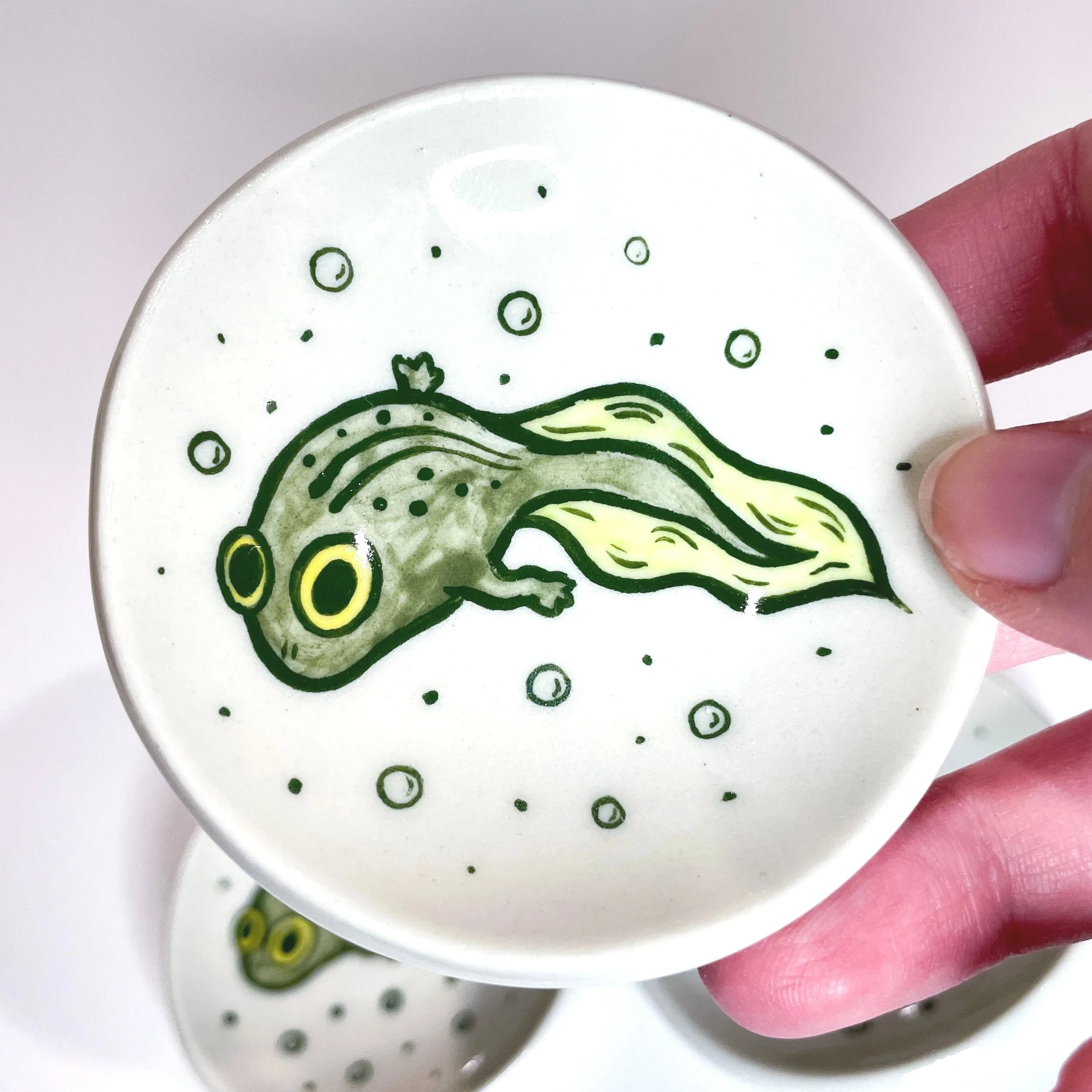 a hand holds up a small trinket dish with a tadpole illustrated on it. there are two other trinket dishes behind it.