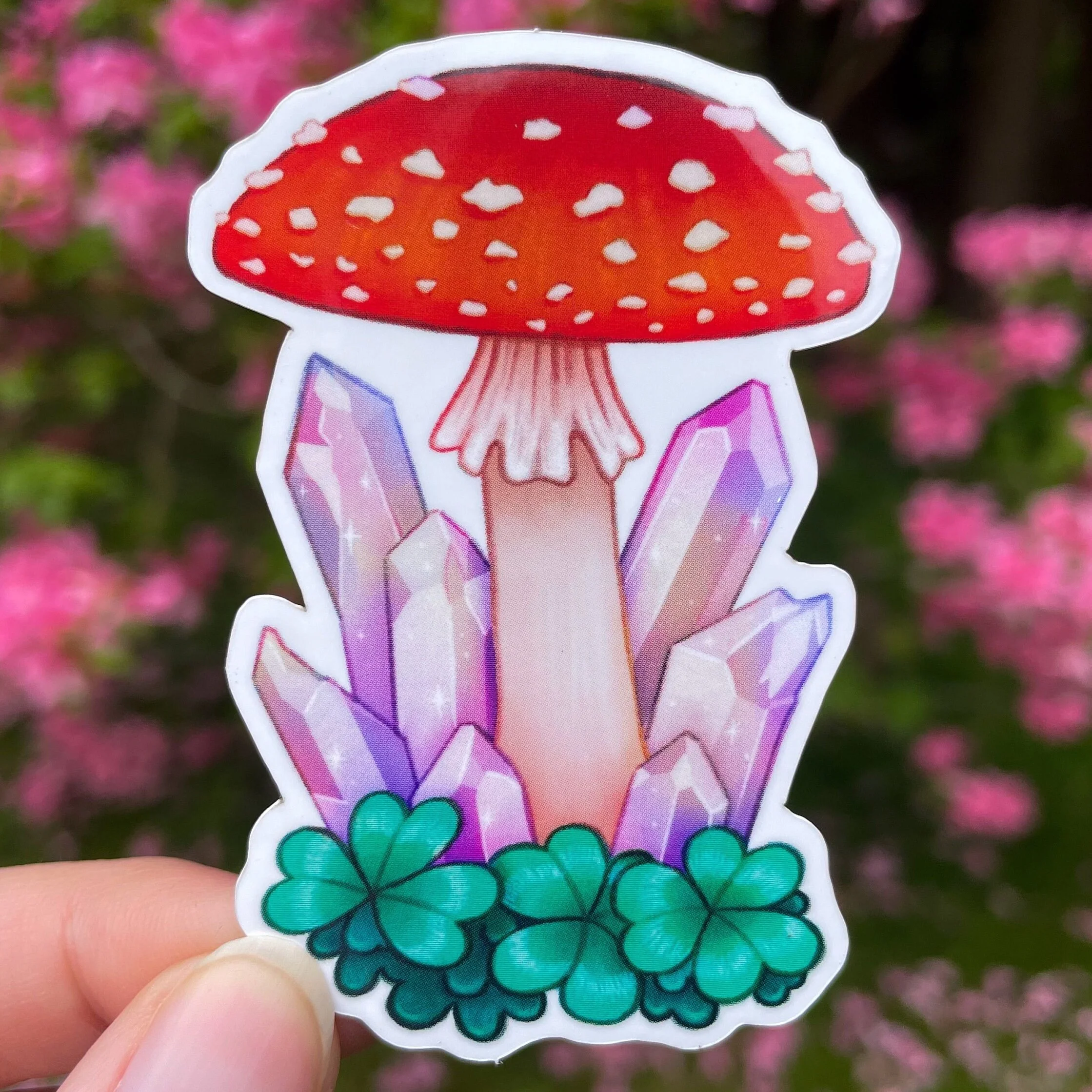 a hand holds up a sticker depicting a red amanita mushroom with crystals and clovers