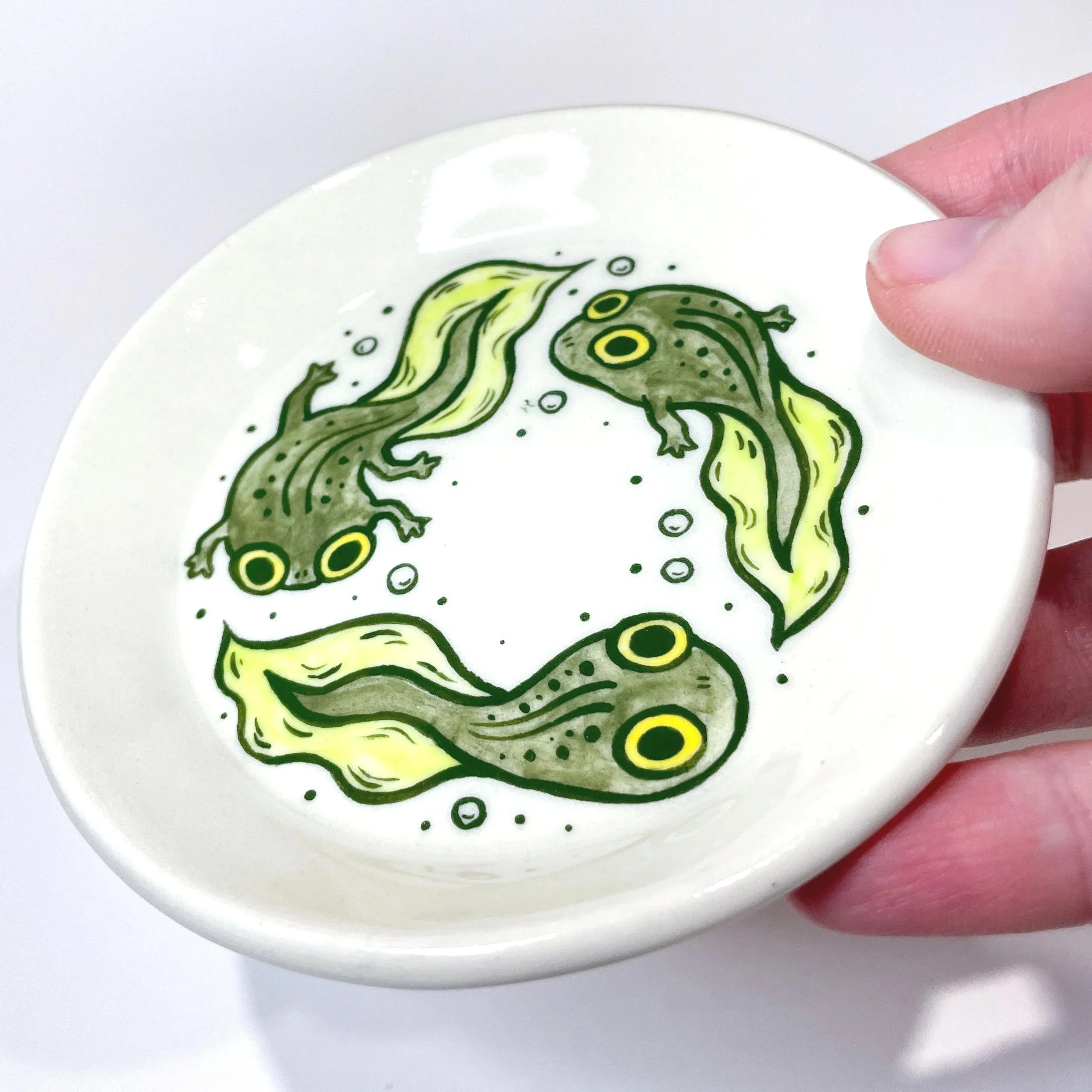 a hand holds up a small trinket dish with three tadpoles illustrated on it.