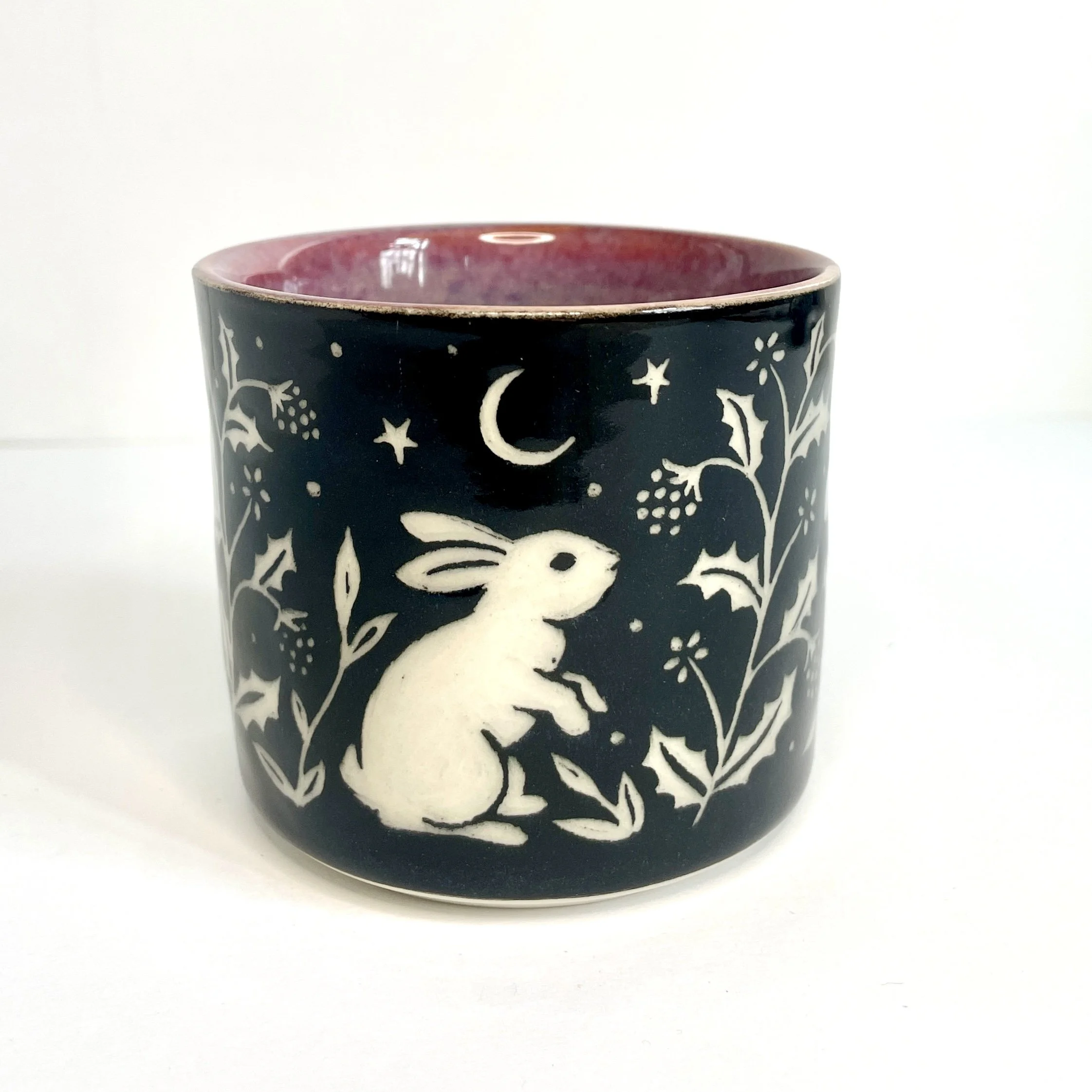 a ceramic cup. The exterior is black and has white bunnies, stars, and leaves. The interior is pink.