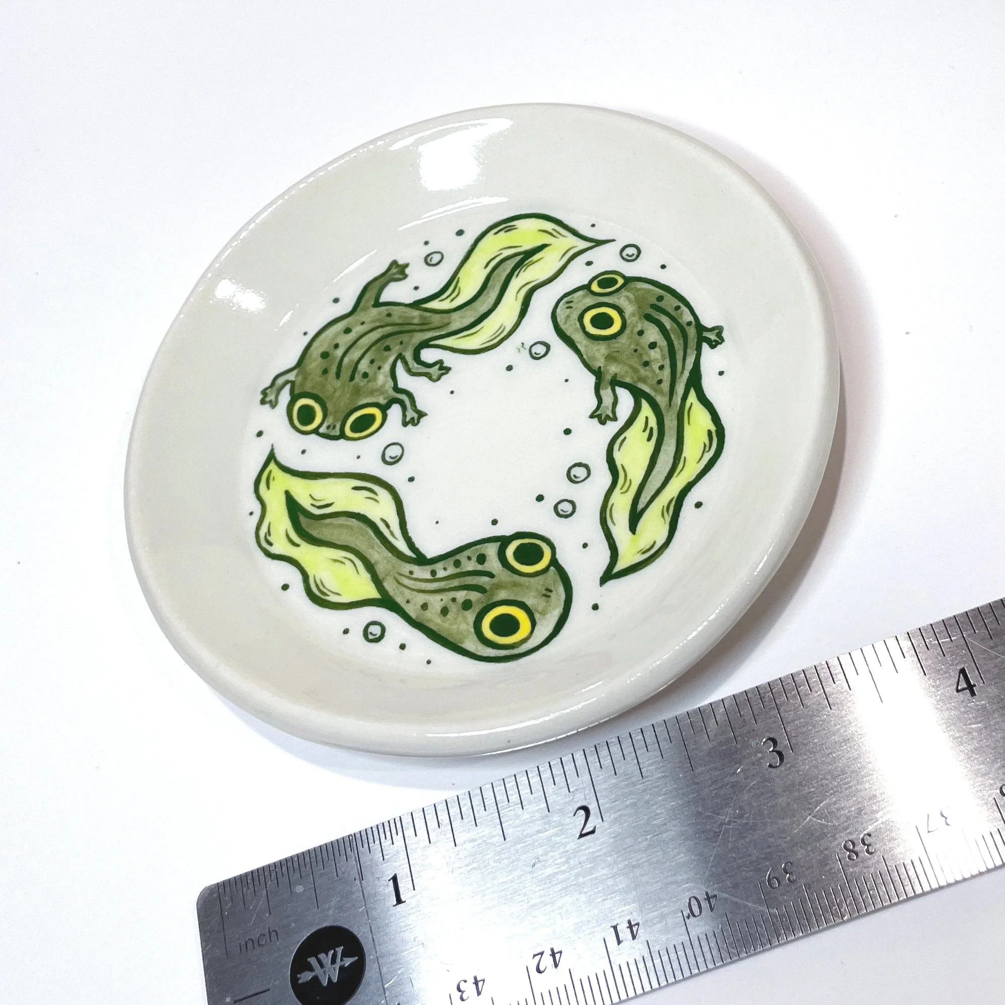 a small trinket dish with three tadpoles illustrated on it next to a ruler showing it is four inches wide.