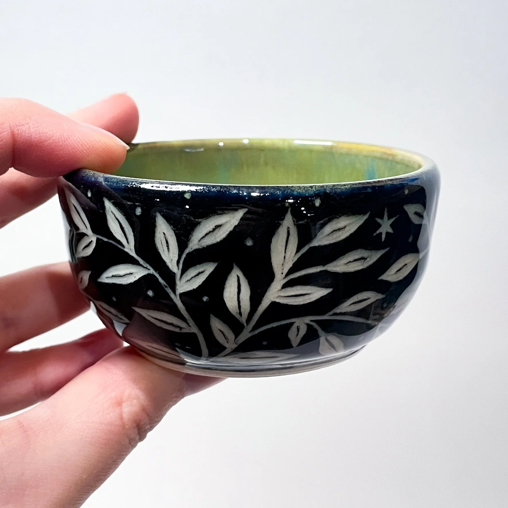A hand holds up a small, black, ceramic trinket bowl with white leaves and stars wrapping around the exterior. The interior is green.