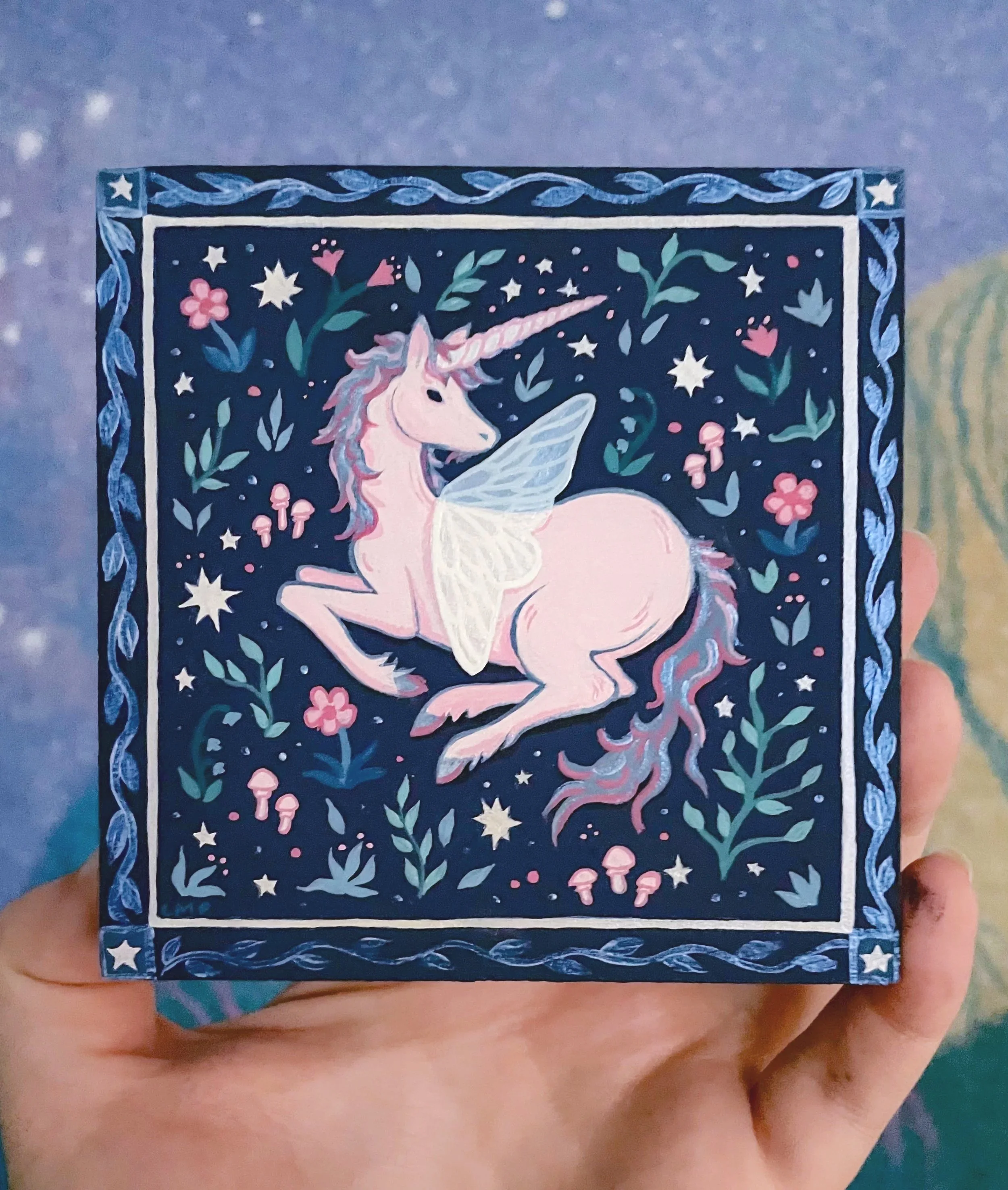 a hand holding up an original painting of a unicorn in a flower field