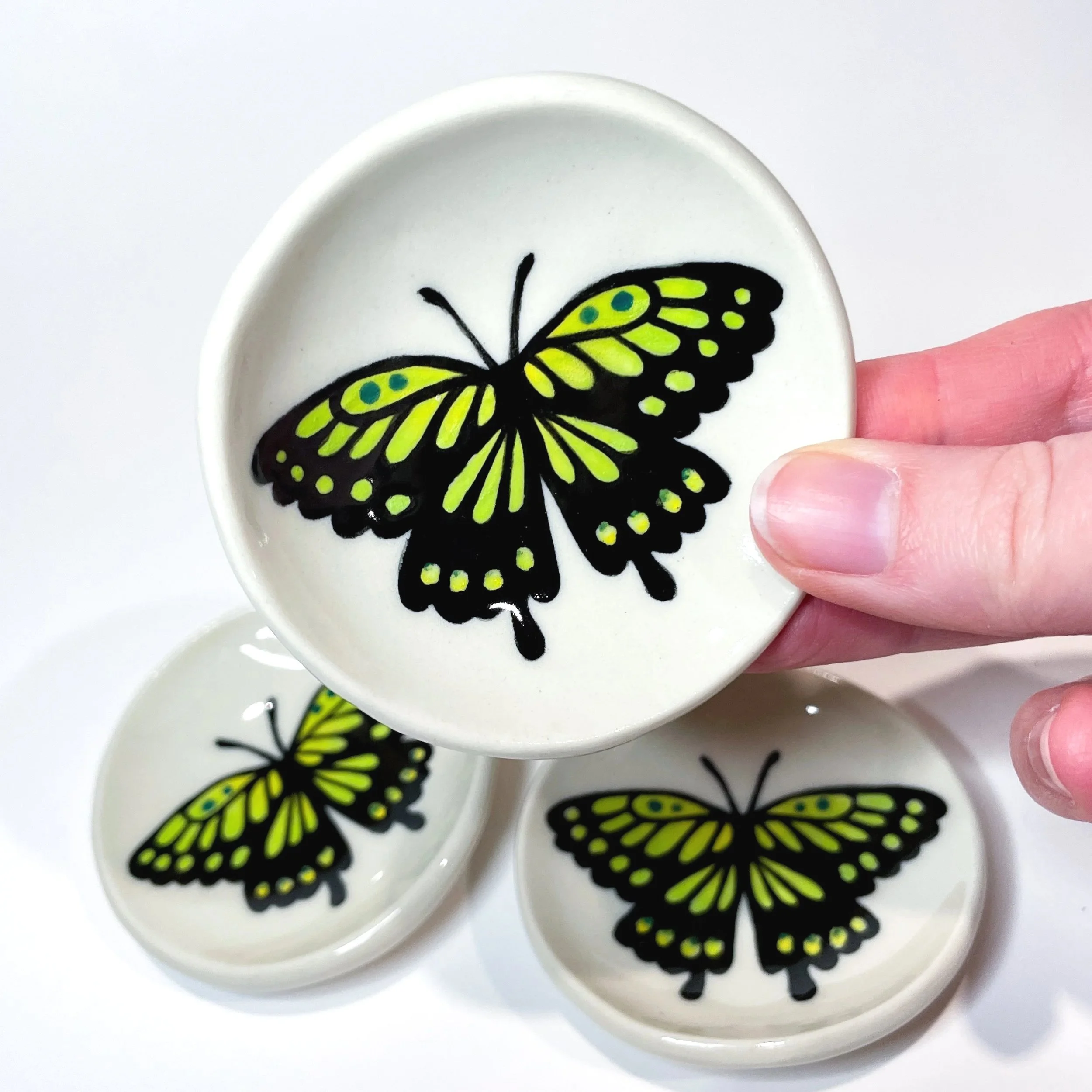 a hand holds up a small ceramic dish with a green and black butterfly on it. there are two more of that dish in the background.