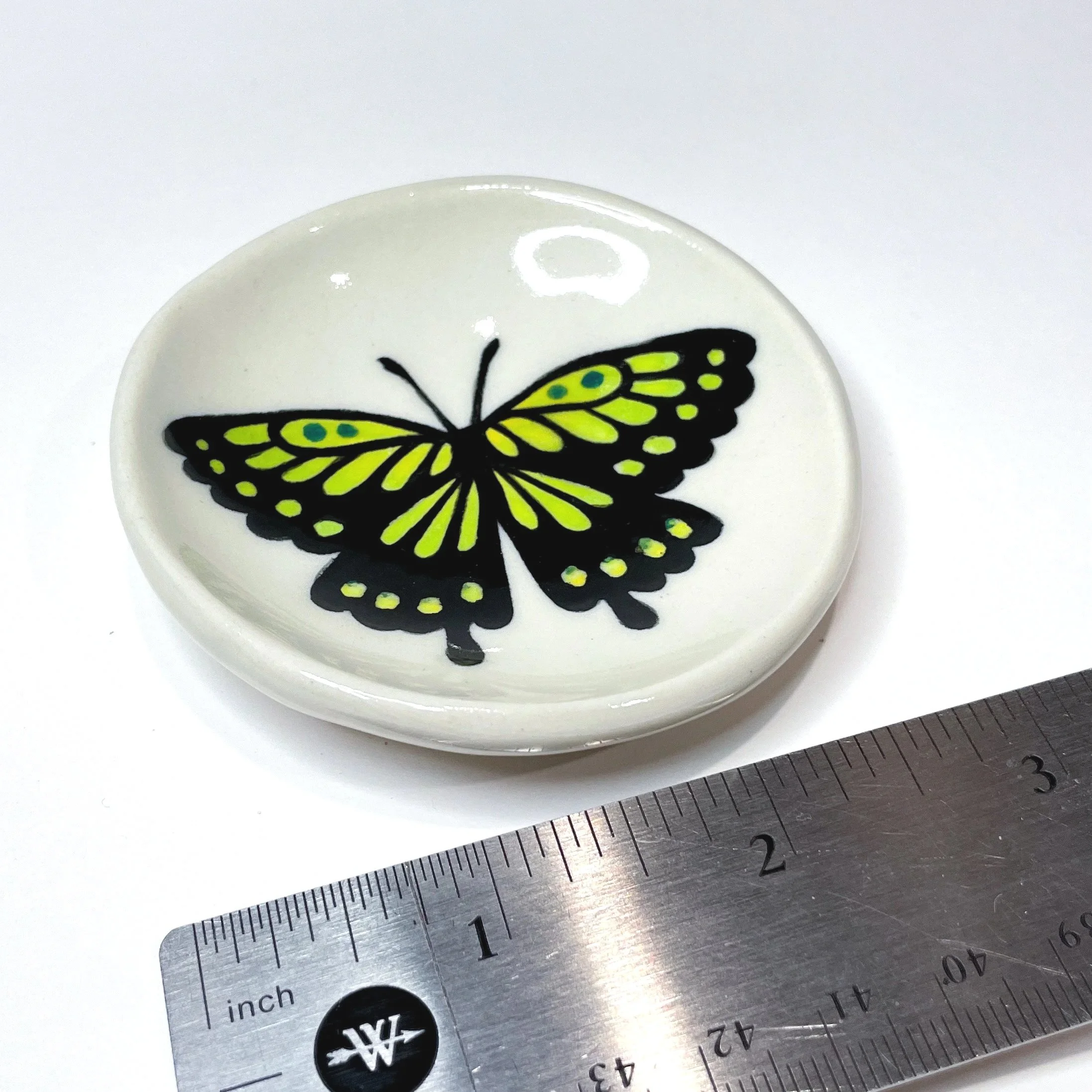 a small ceramic dish with a green and black butterfly on it next to a ruler showing it is three inches wide.
