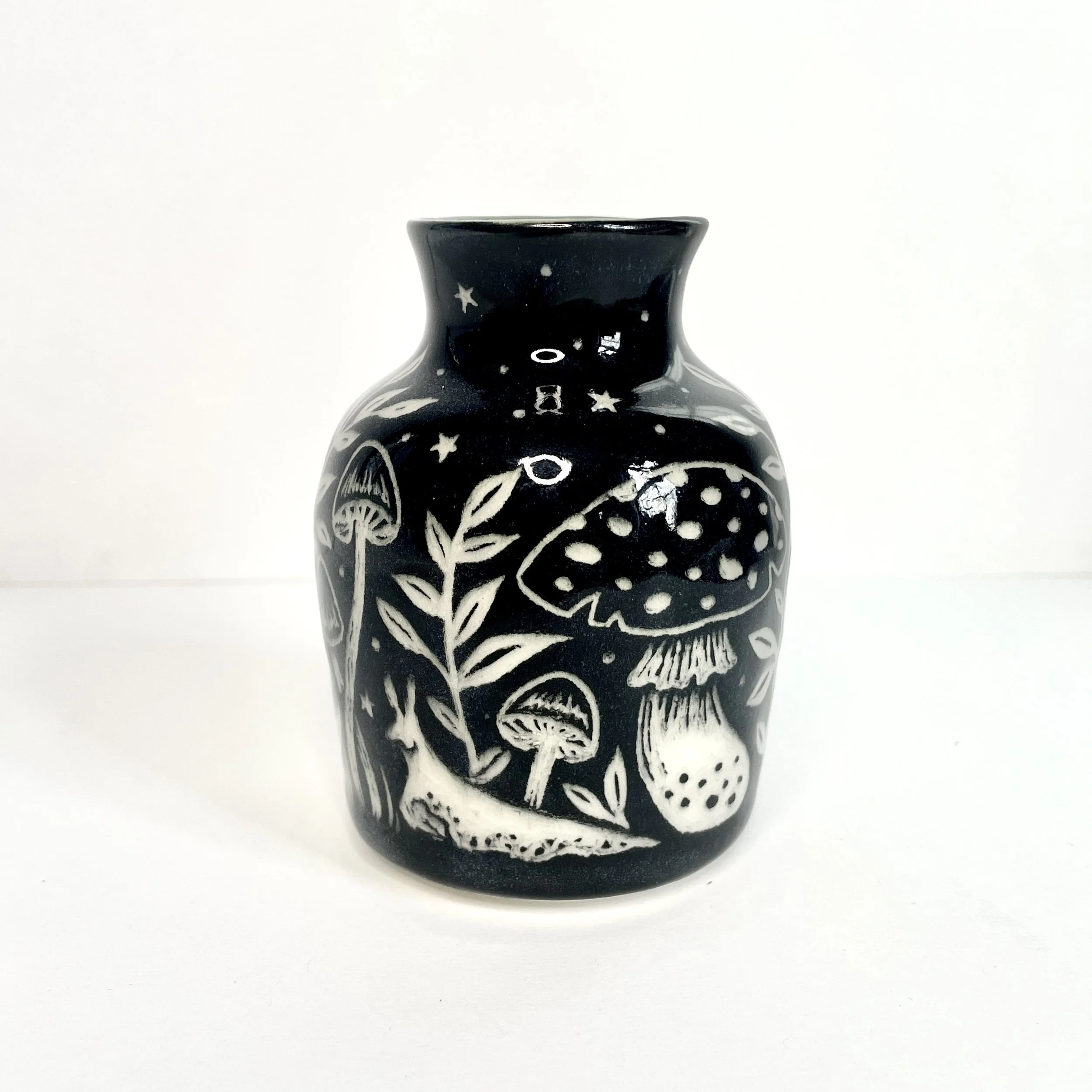 a ceramic bud vase. the exterior is black and has white snails, mushrooms and leaves. the interior has green around the rim.