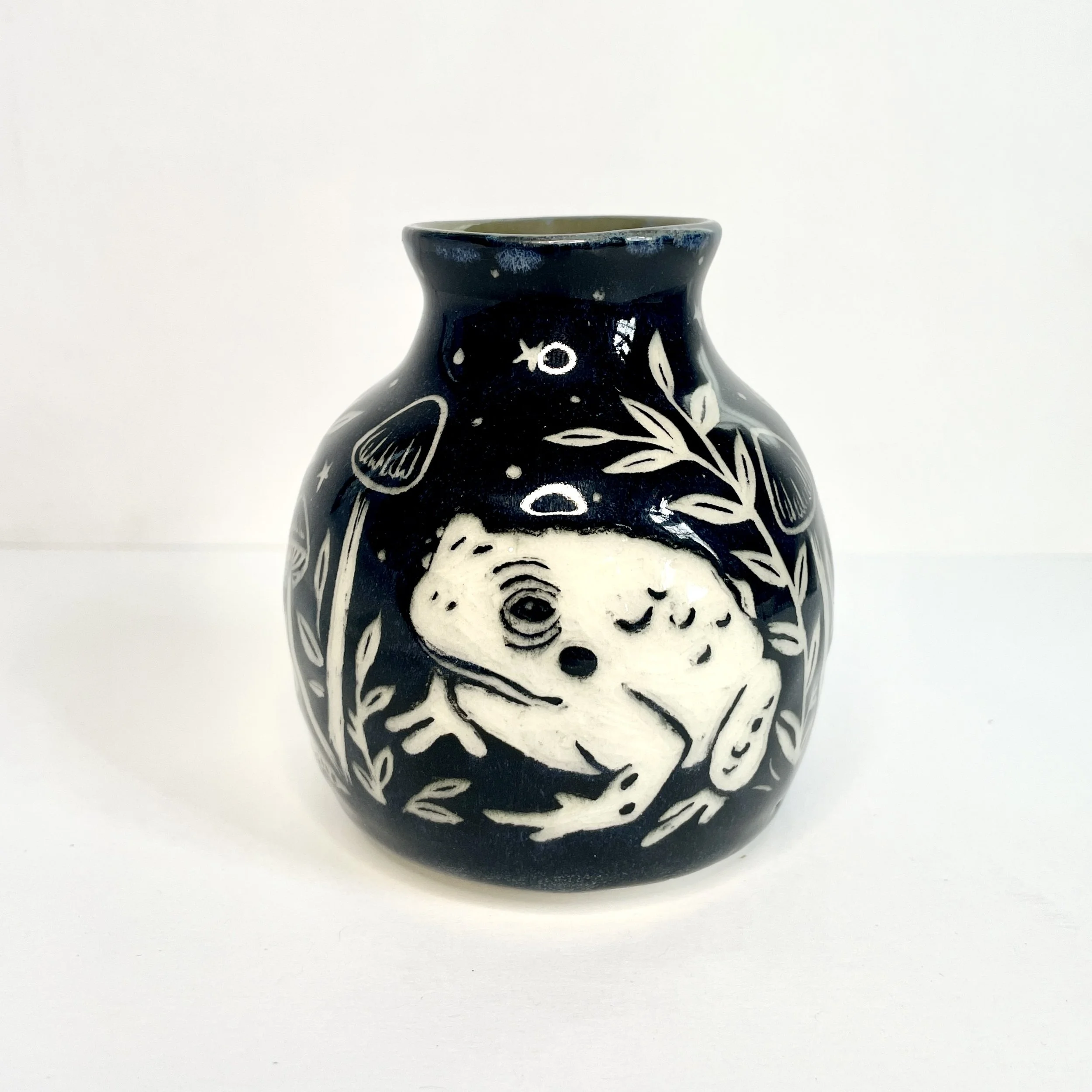 a small ceramic bud vase. the exterior is black and has white toads and leaves. the interior has green around the rim.