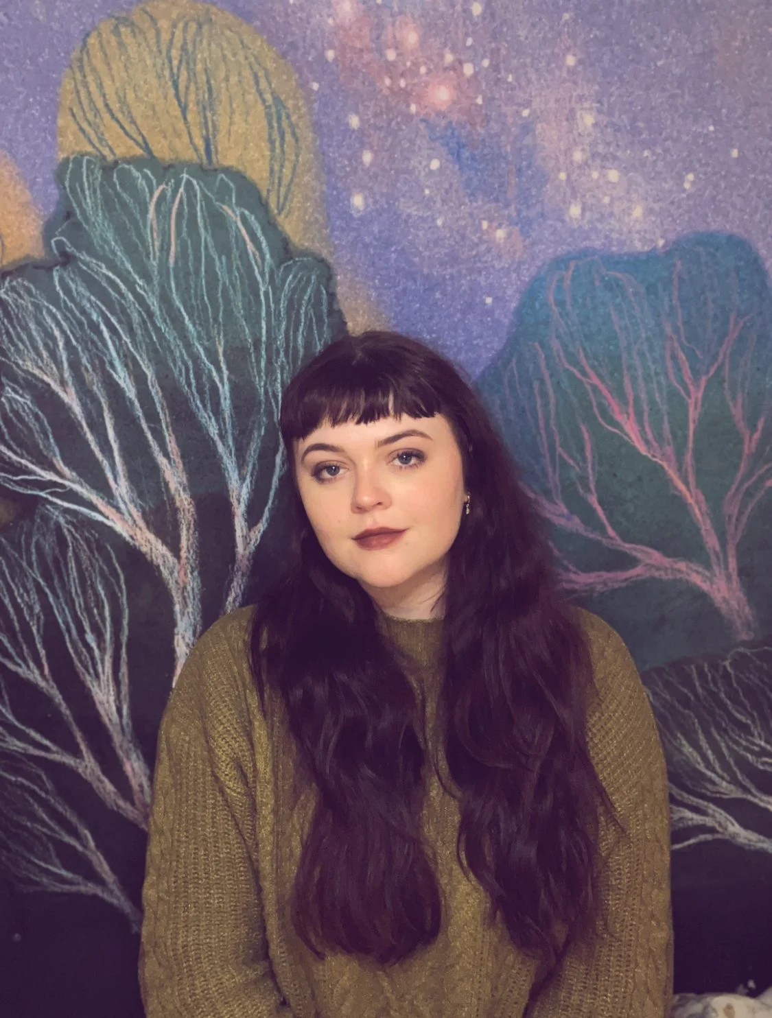 a pale girl with long dark hair wearing a green sweater in front of an illustrated forest background.