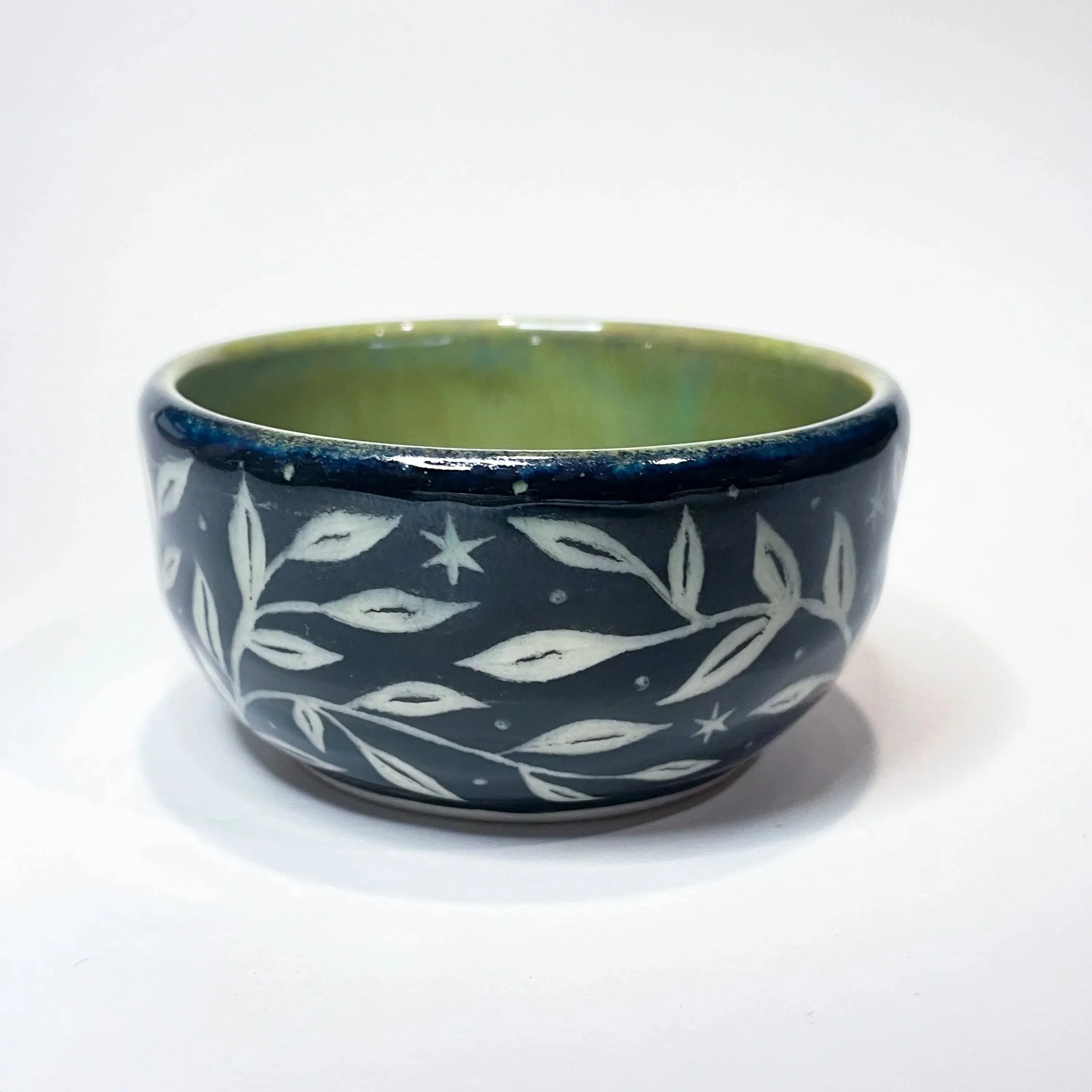 A small, black, ceramic trinket bowl with white leaves and stars wrapping around the exterior. The interior is green.