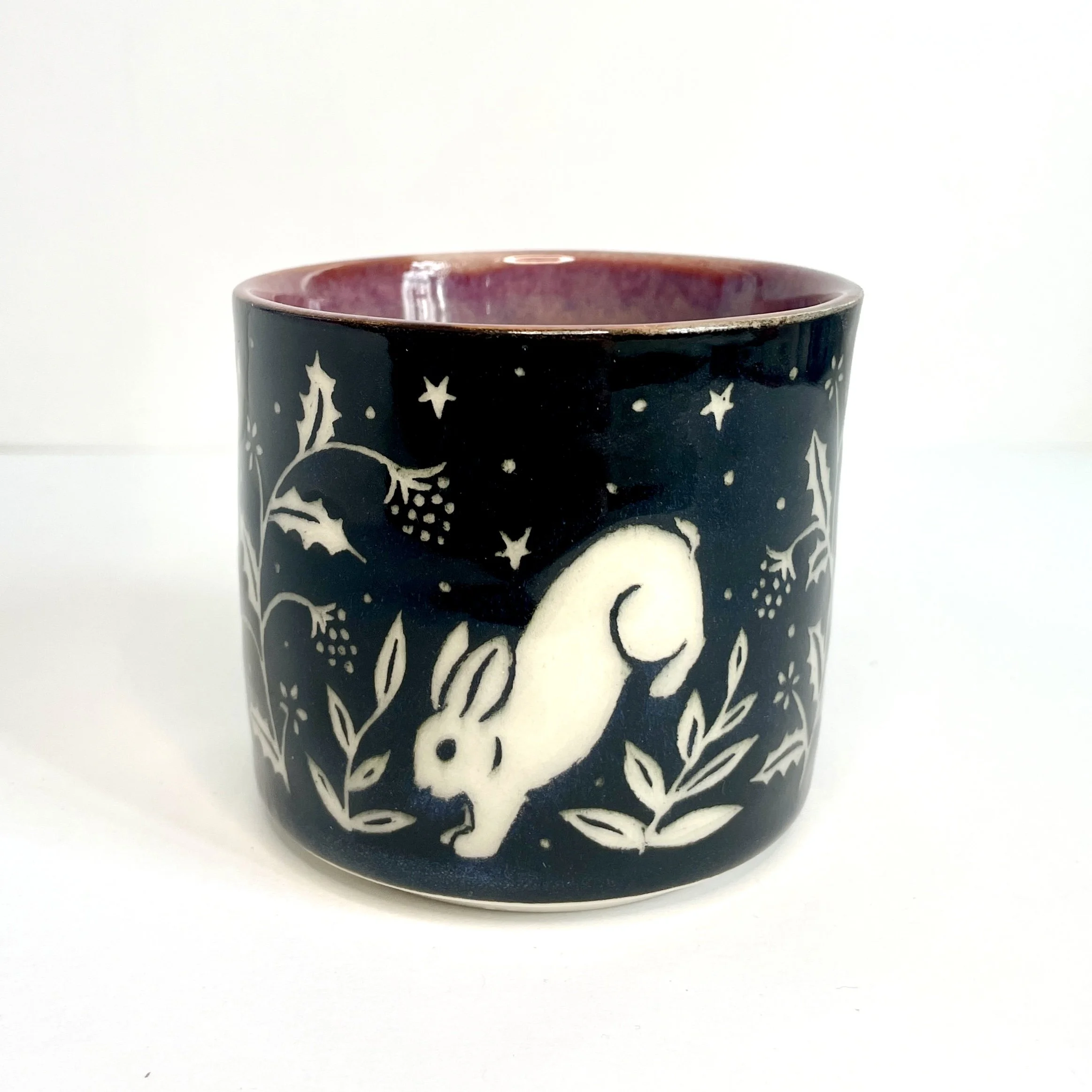 a ceramic cup. The exterior is black and has white bunnies, stars, and leaves. The interior is pink.