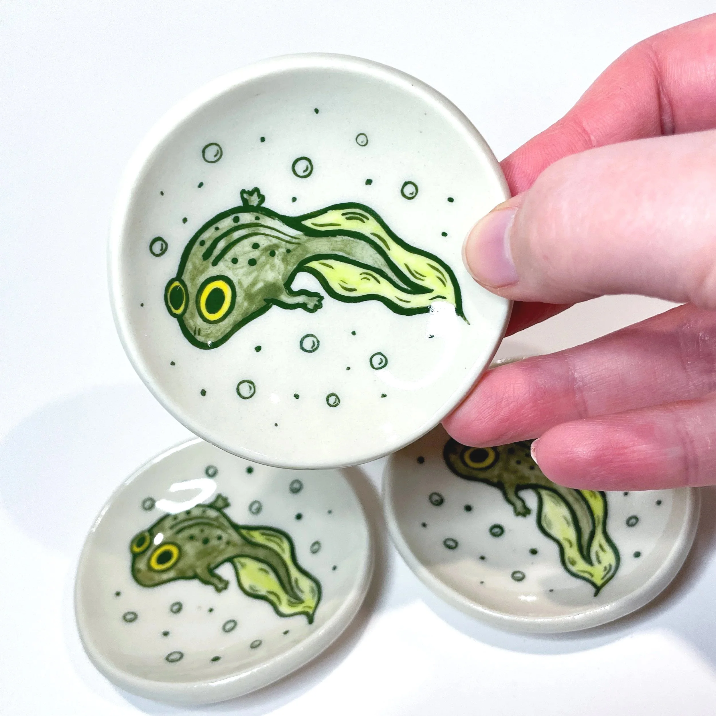 a hand holds up a small trinket dish with a tadpole illustrated on it. there are two other trinket dishes behind it.