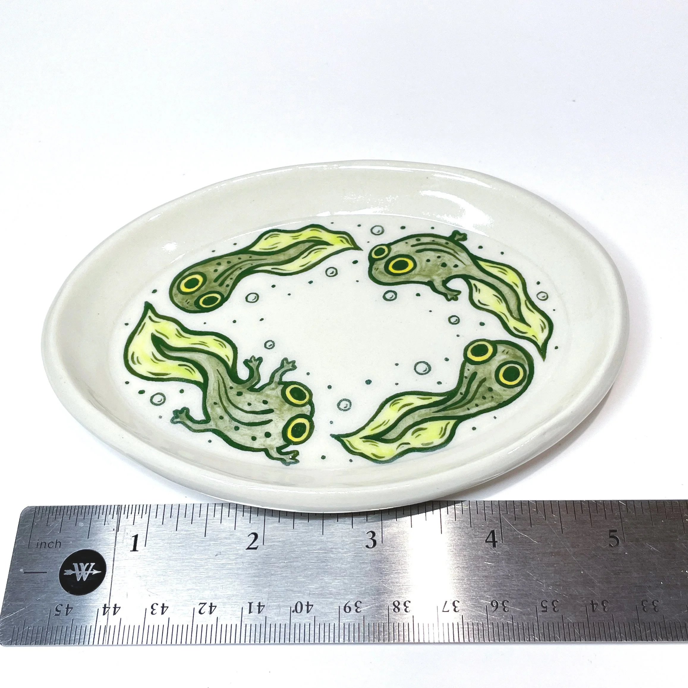 an oval trinket dish with four tadpoles illustrated on it next to a ruler showing it is five inches wide.