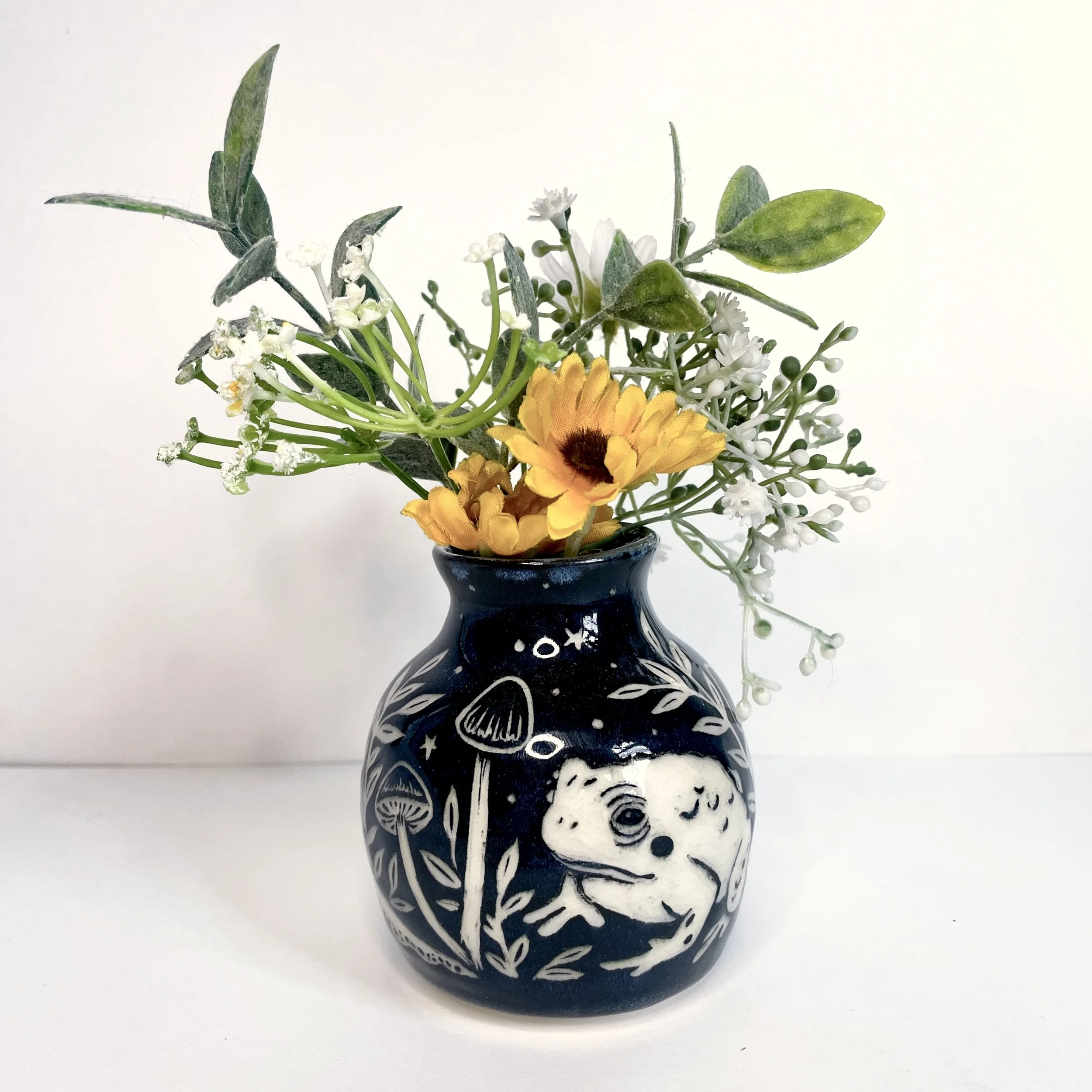 a small ceramic bud vase. the exterior is black and has white toads and leaves. the interior has green around the rim. this product photo includes flowers and greenery inside the vase.