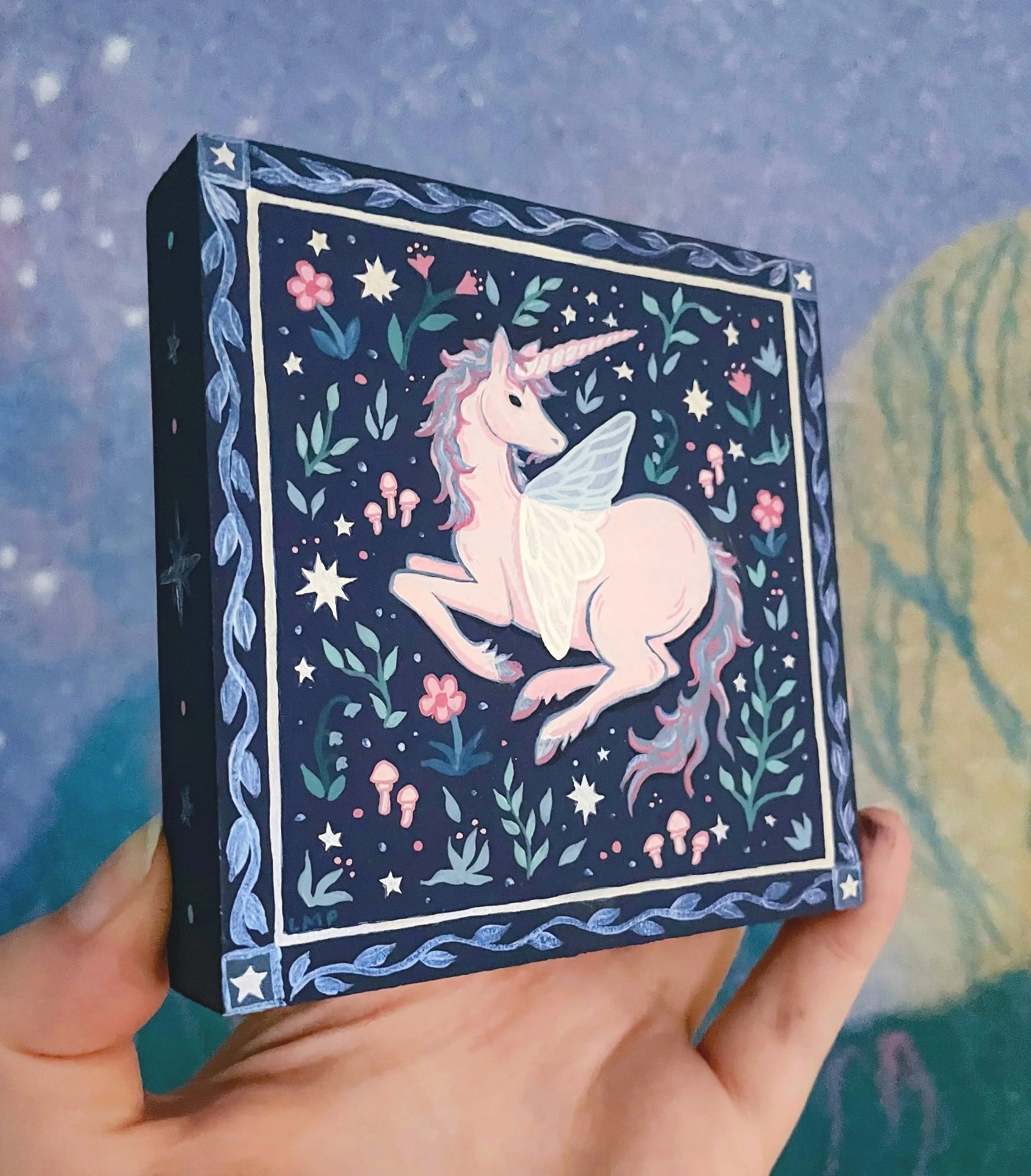 a hand holding up an original painting of a unicorn in a flower field