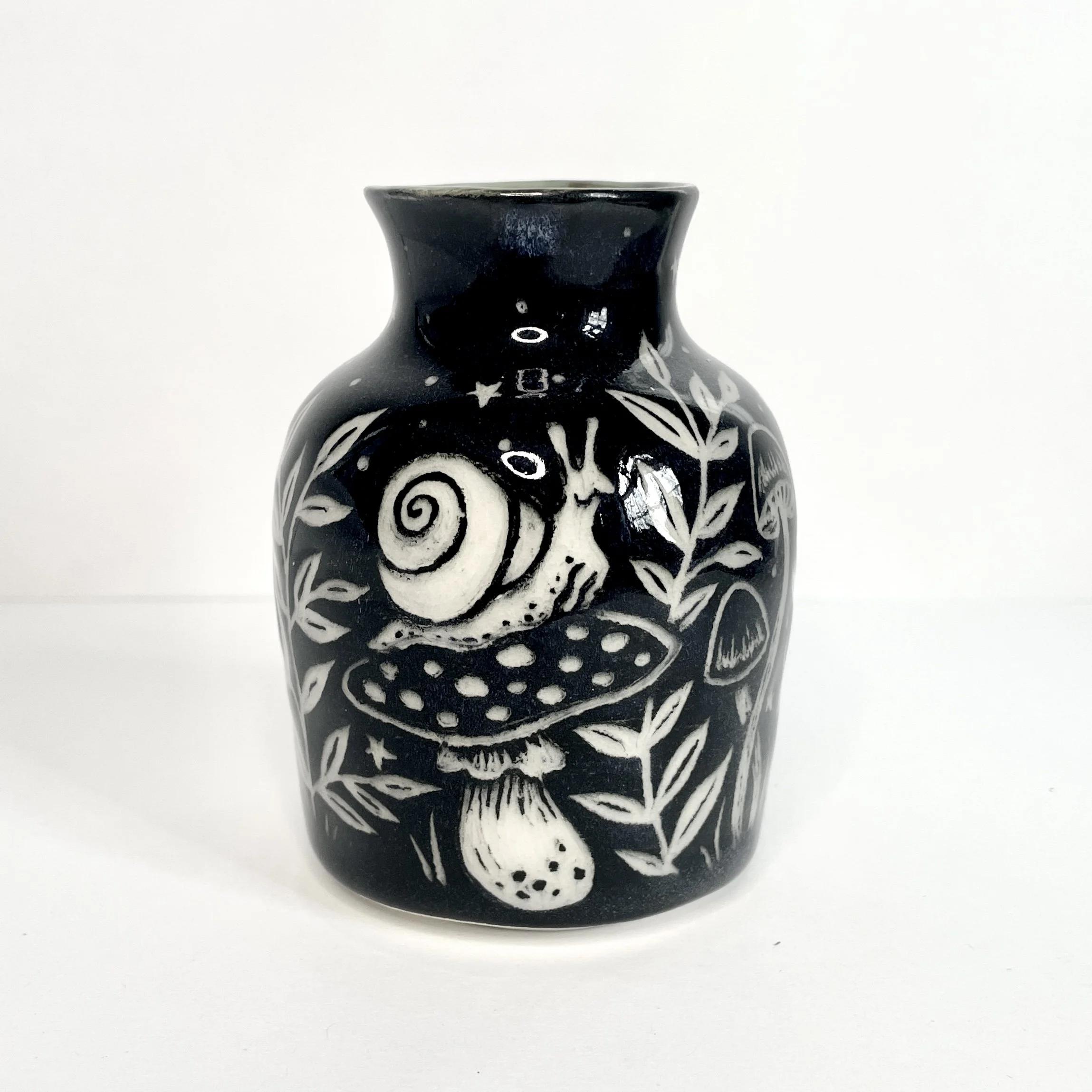 a ceramic bud vase. the exterior is black and has white snails, mushrooms and leaves. the interior has green around the rim.