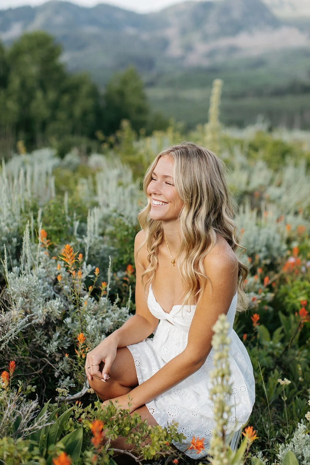 High school senior sitting in a wildflower field of orange flowers, laughing to the side and wearing a white dress. Hael Studio. Park City Utah Senior Photos. Digital and 35mm film photography.