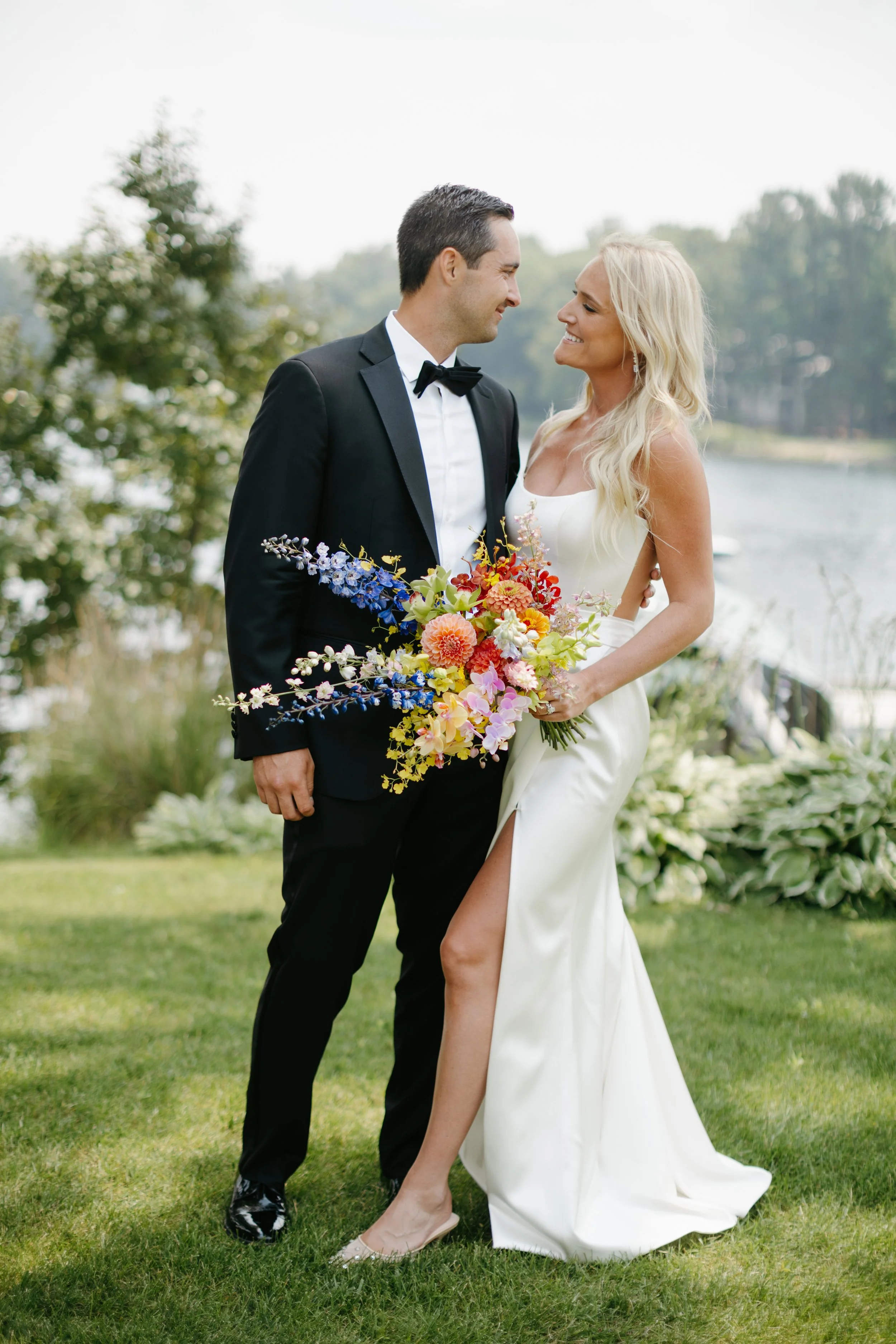 Bride and groom standing on grass, looking at each other while the bride holds a bouquet of rainbow colored flowers. Private Lake Minnetonka Backyard Wedding. Digital and 35mm film Minnesota Wedding Photographer.