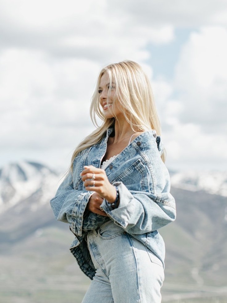 High school senior wearing a denim jacket and jeans with blue and white snow-covered mountains in the background. Park City Utah Senior Photos. Digital and 35mm film photography.