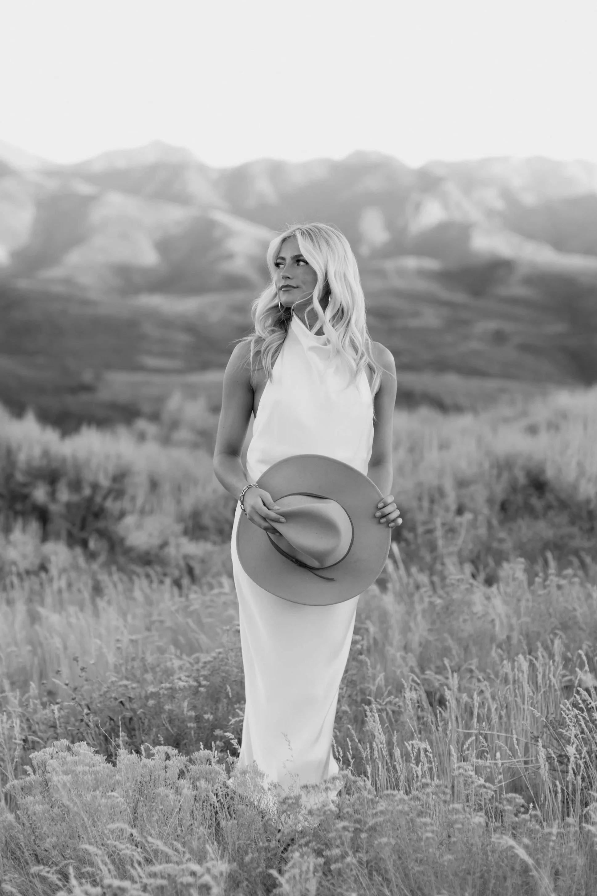 Woman looking to her right, while holding a cowboy hat and standing in a field with the mountains behind her. Park City Utah Wedding Photographer. Hael Studio. Leah Mae Photography.
