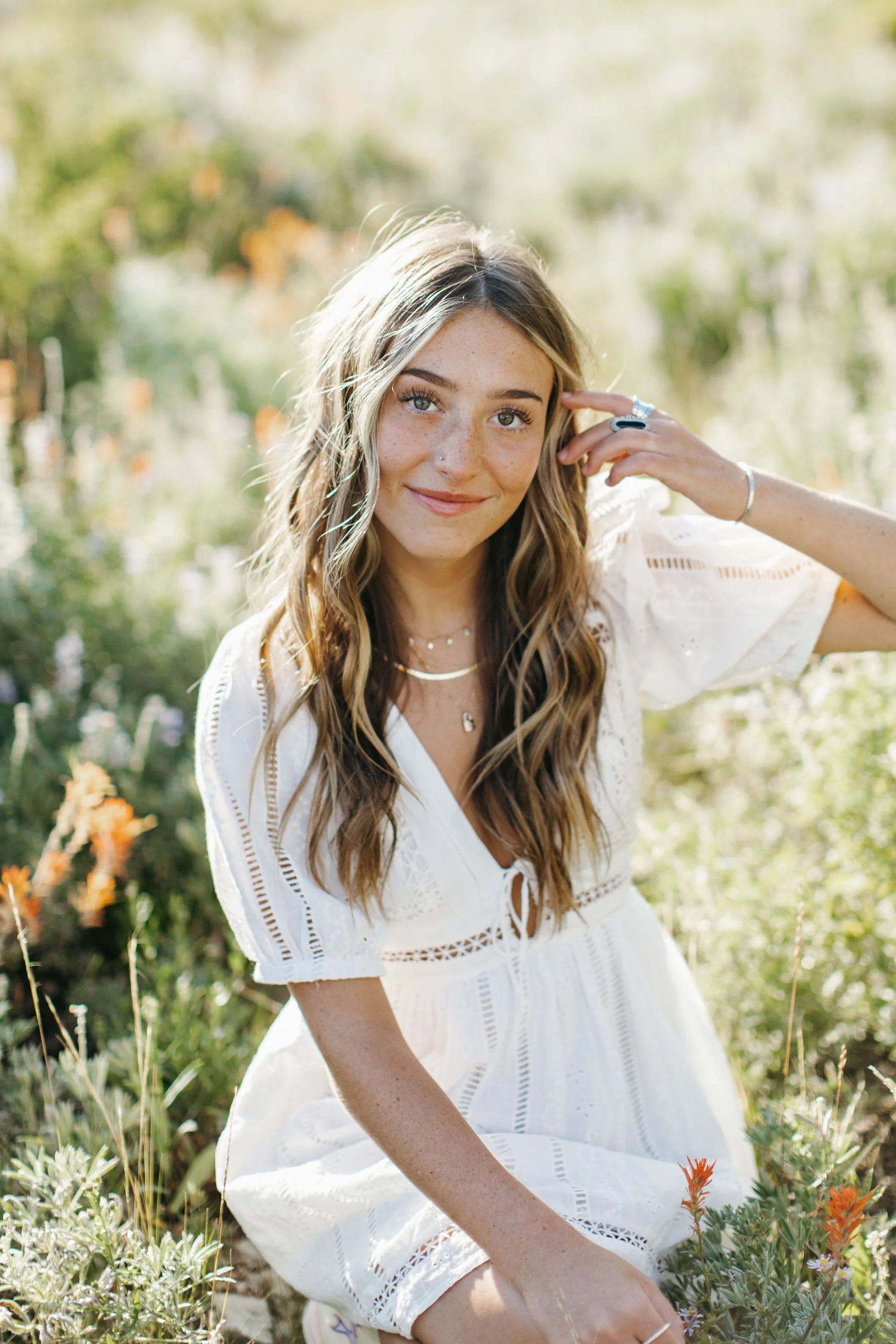 High school senior sitting in a wildflower field, holding one hand up to her face and wearing a white dress. Park City Utah Senior Photos. Digital and 35mm film photography.