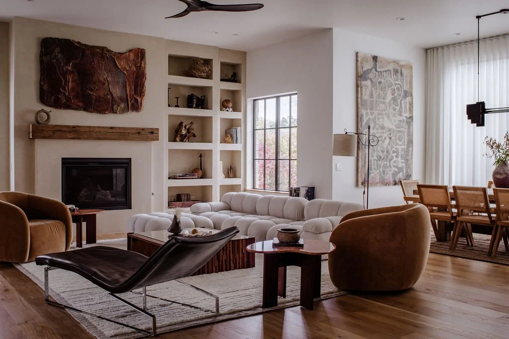 Living room with white sofa, brown armchairs, black lounge chair, wooden coffee table, fireplace, built-in shelving, large window, abstract wall art, dining area with wooden table and chairs, and decorative plants.