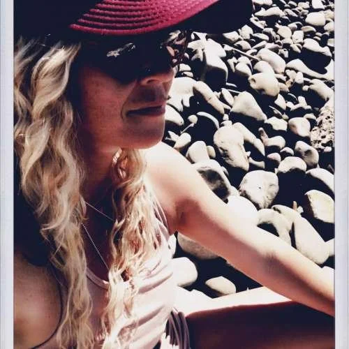 Woman with long curly blonde hair, sunglasses, and a cap, smiling outdoors on a trail surrounded by greenery.
