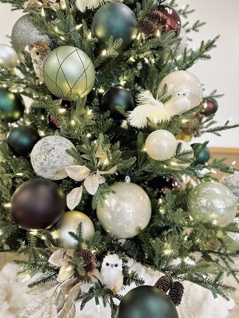 Close-up of a decorated Christmas tree with ornaments, including a yellowish-green ball with black glittered lines, a creamy white ball, and a white bird figurine with feathers, illuminated by small yellow lights.