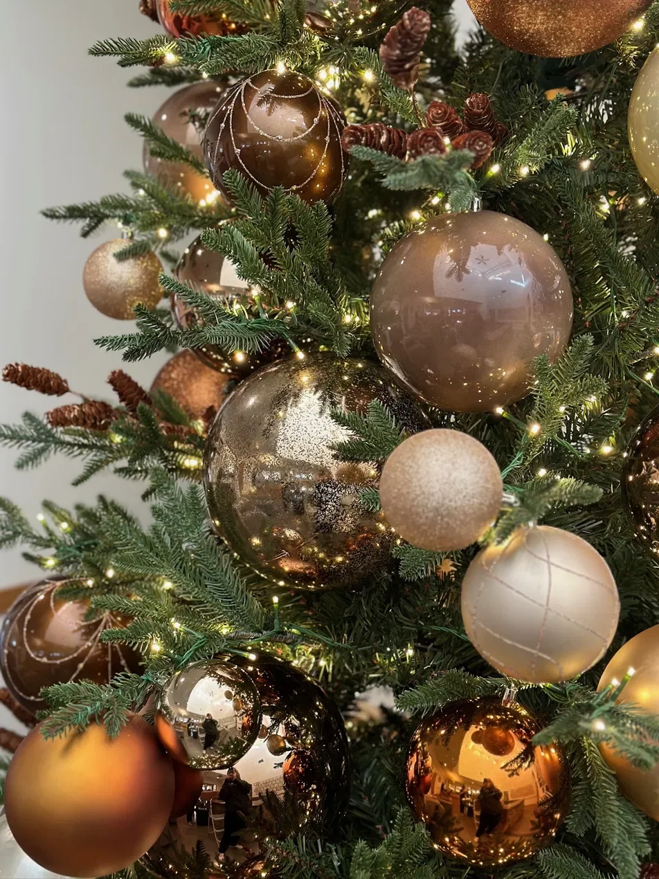Close-up of a decorated Christmas tree with gold and copper ornaments, including shiny, matte, and glittery balls, with warm fairy lights on green pine branches.