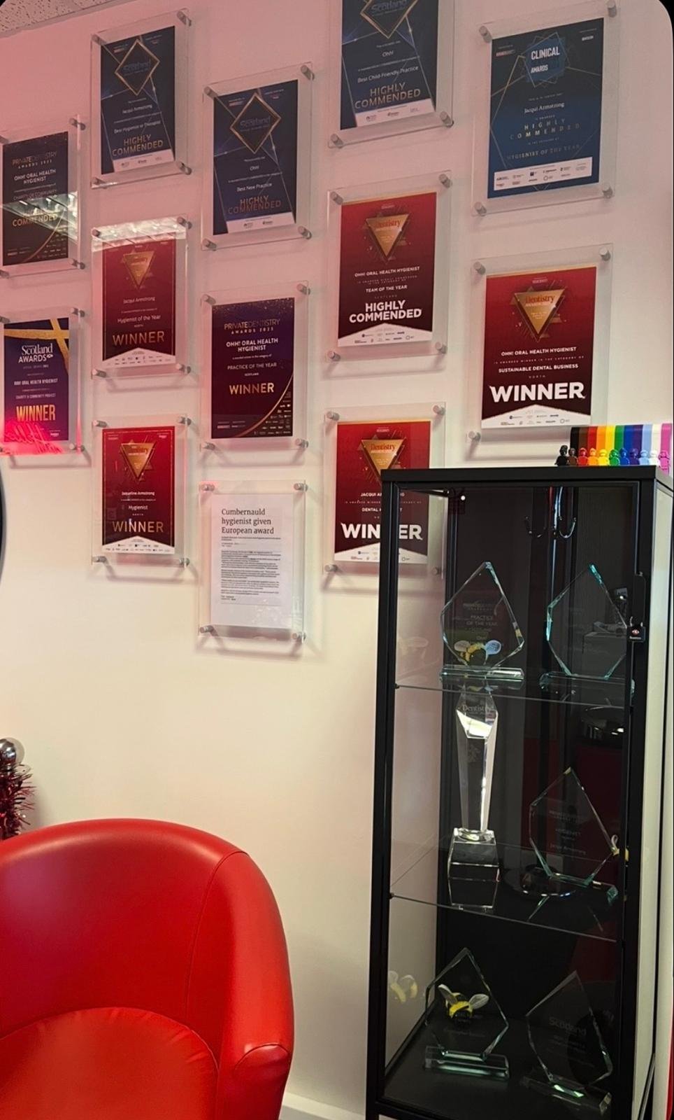Wall with multiple award certificates in glass frames, a black display case with glass awards inside, a red chair, and colorful pens on a table.