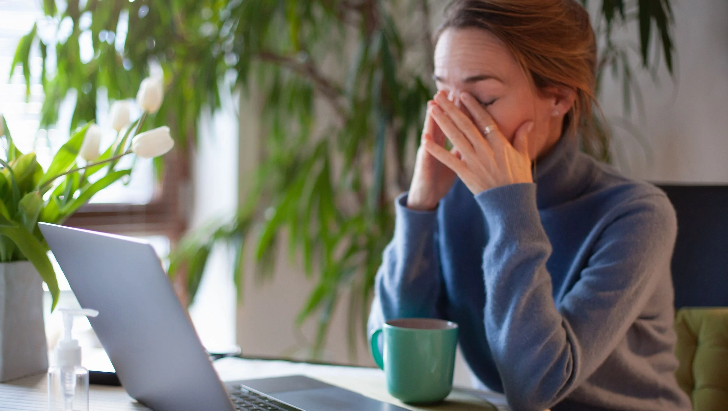 A woman with reddish hair, wearing a gray sweater, sitting at a desk with a laptop, holding her face with her hands, appearing to be emotional or tired, with green plants in the background and a green mug on the desk.