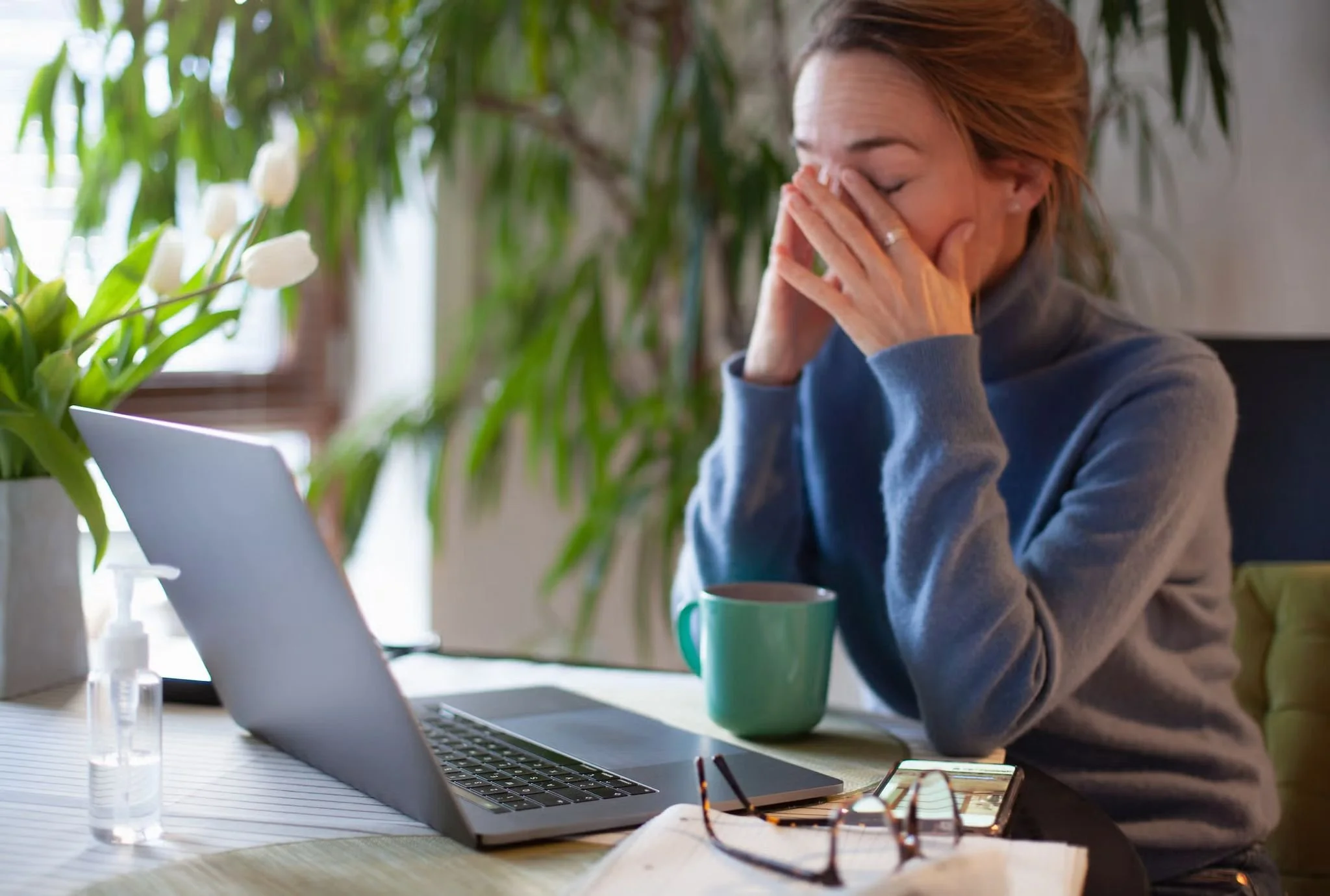 A woman sitting at a desk with a laptop, holding her face with her hands, appearing stressed or tired, with a green mug, glasses, a notebook, and a plant nearby.