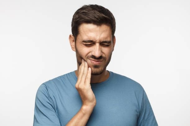 Man holding side of his face in pain or discomfort, grimacing with eyes closed, wearing a blue t-shirt against a plain white background.