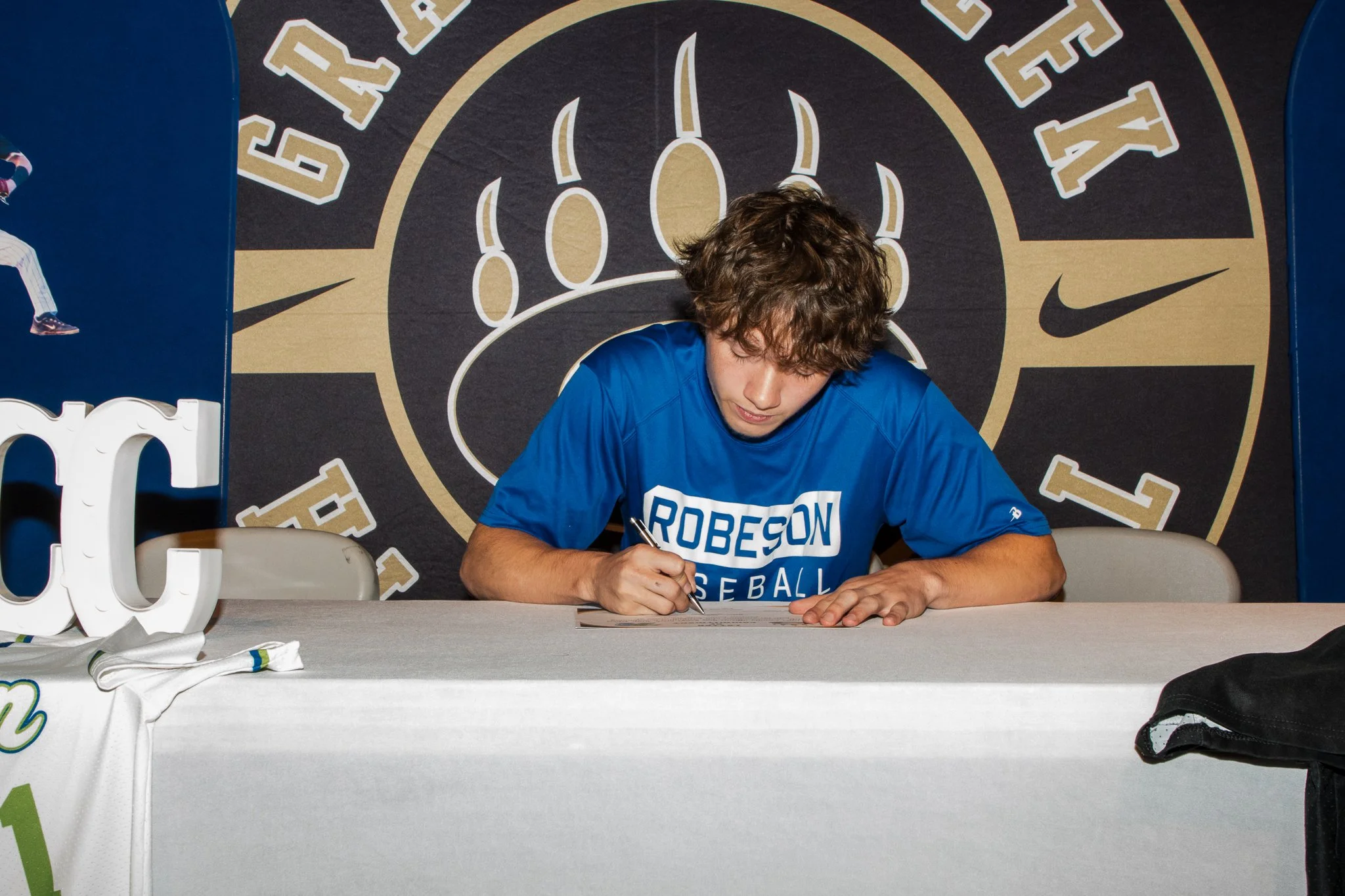 Person wearing a 'Methodist' T-shirt signing a paper at a table with a panther statue on it, against a backdrop displaying 'Hills School' logos.