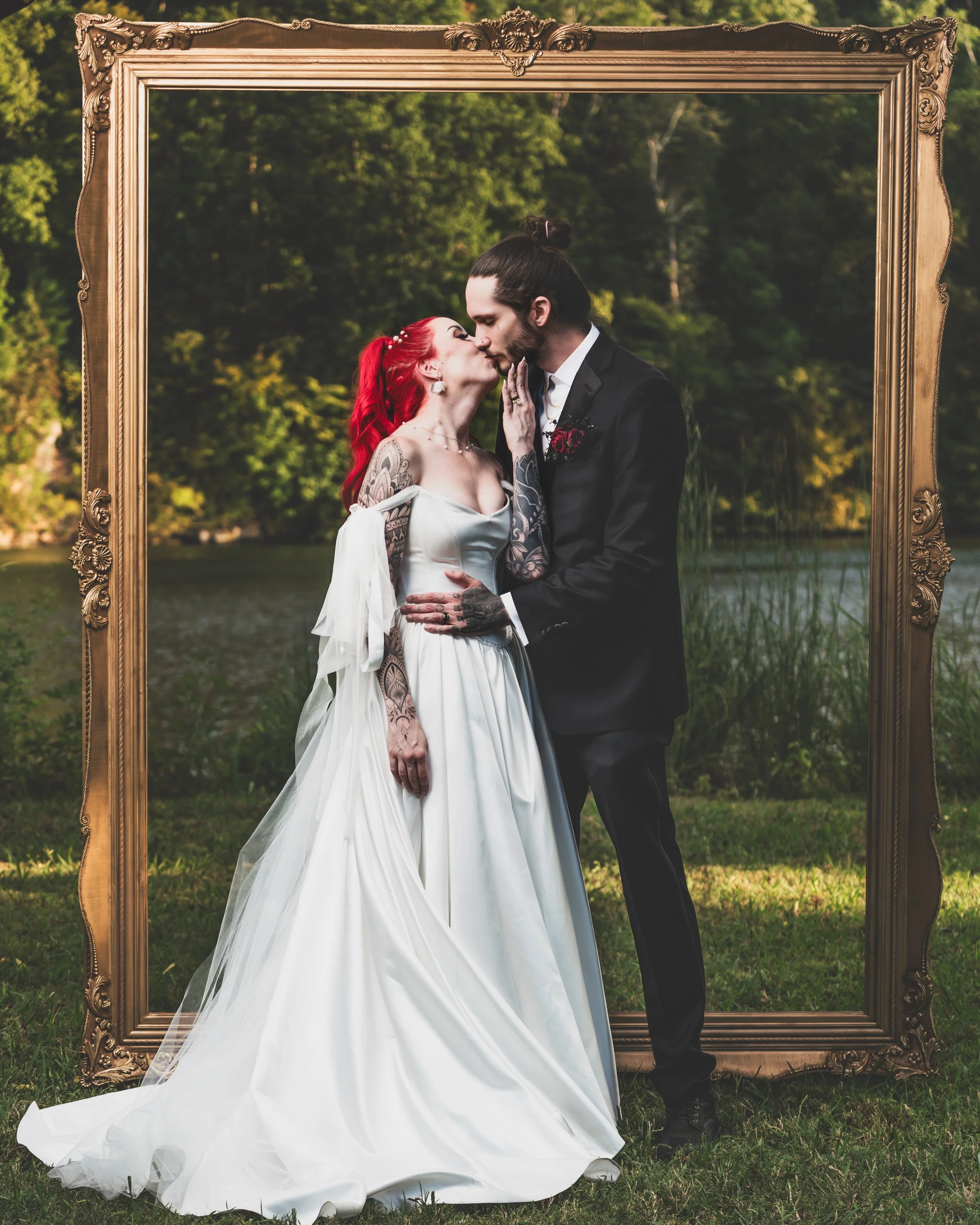 A bride and groom sharing a kiss outdoors with a decorative gold frame around them, lush greenery and a lake in the background.
