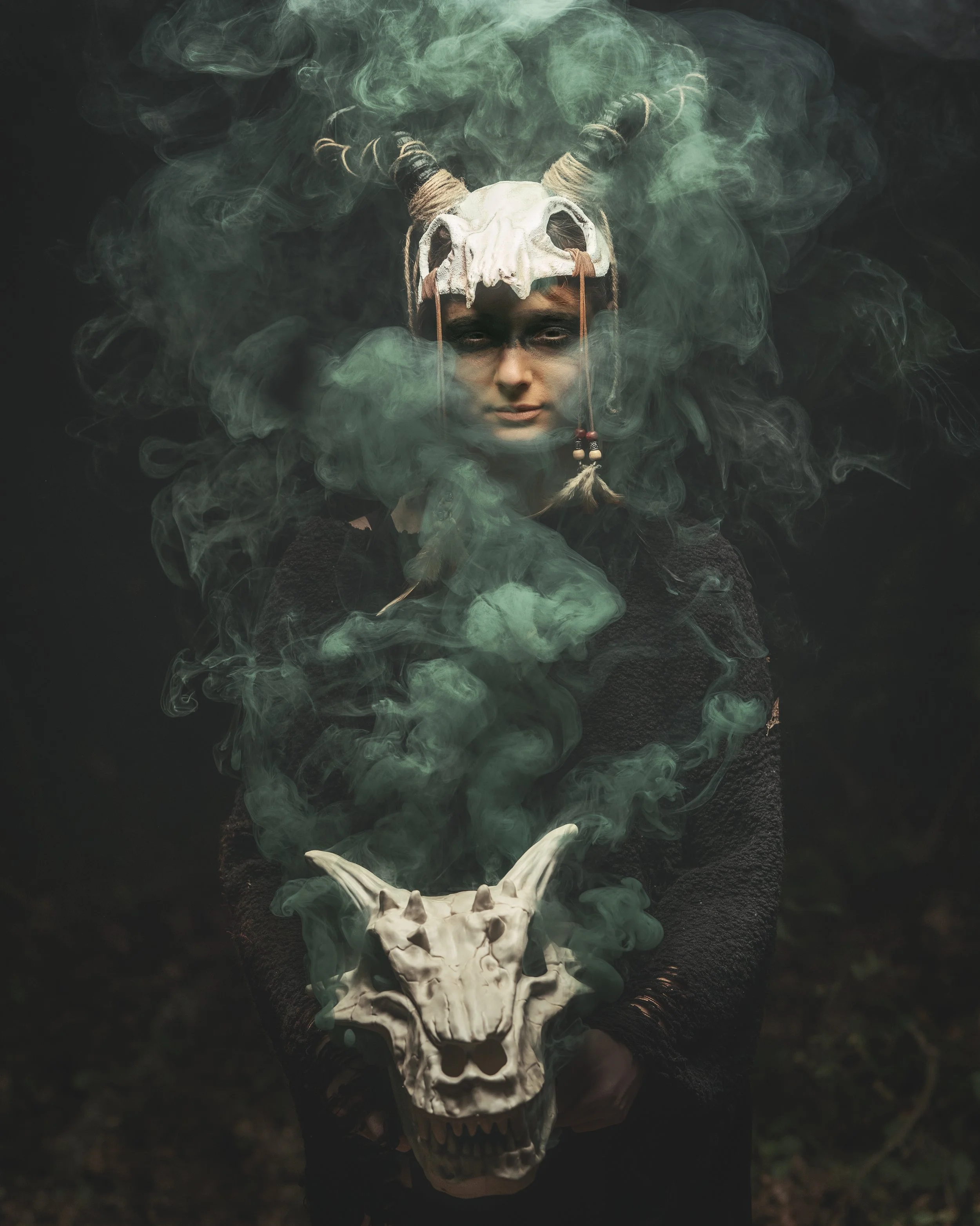 Person wearing a skull and horns mask with green smoke and holding a horned skull in front of a dark background