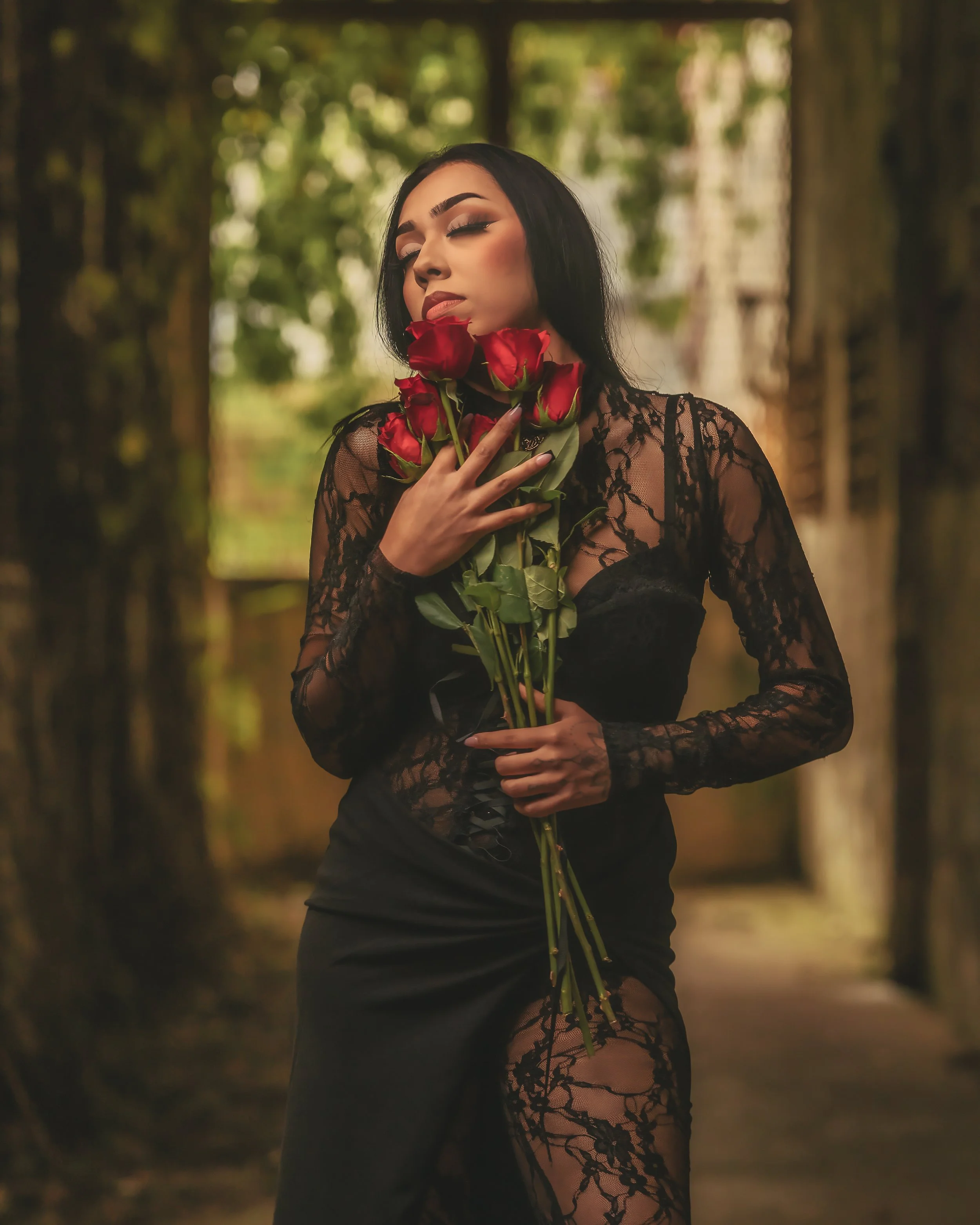 A woman in a black lace dress holding a bouquet of red roses in an outdoor wooded area.