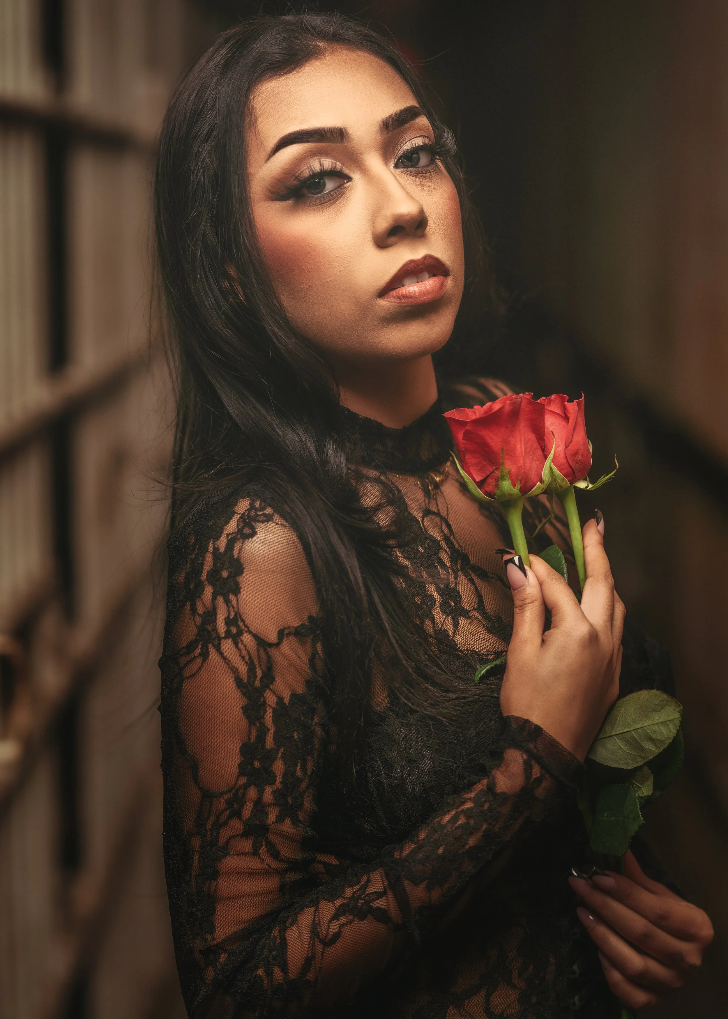 A woman with dark hair and makeup holding a red rose in her hand, wearing a black lace dress, in front of a background with shelves of books.