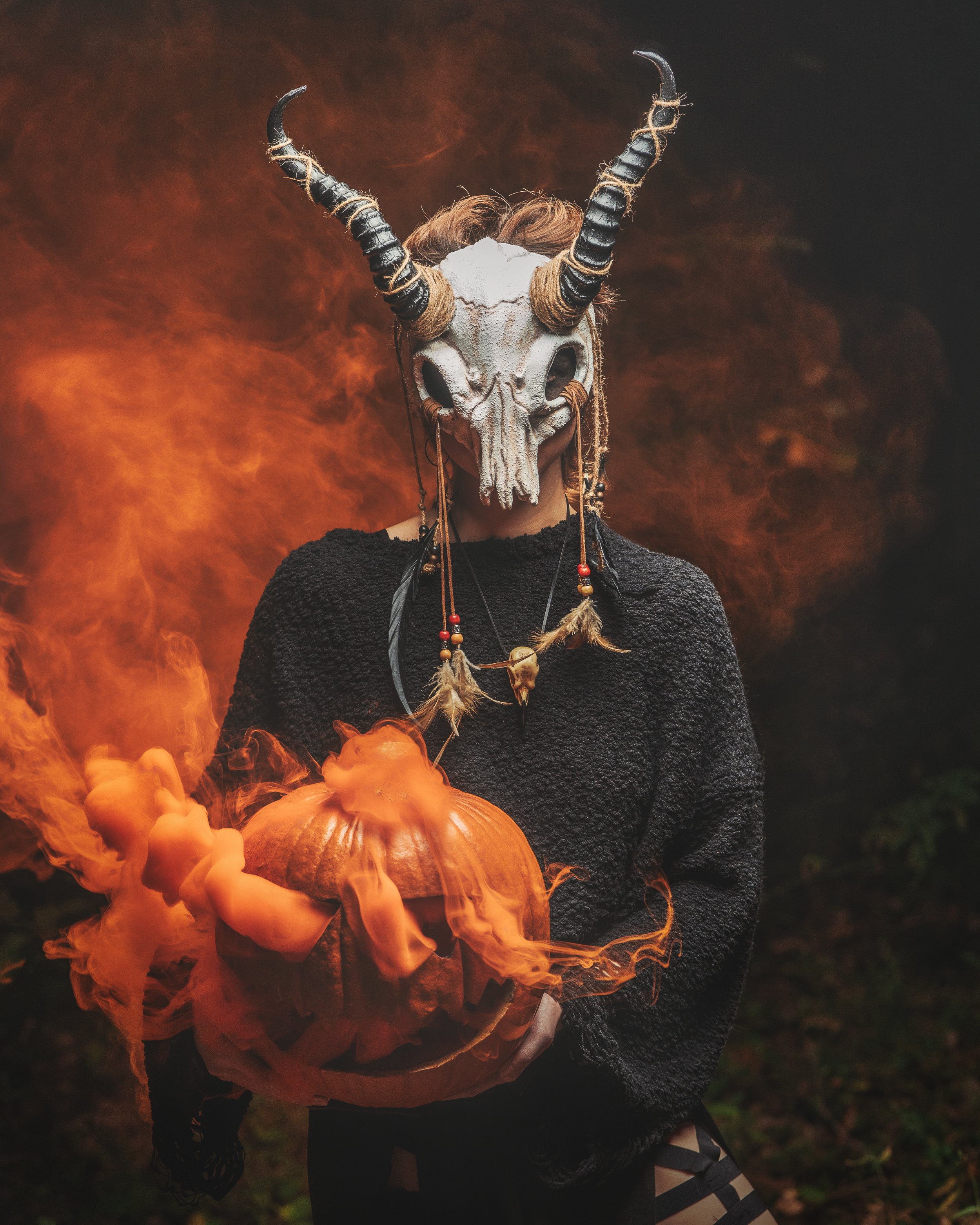 Person dressed in a black textured sweater wearing a goat skull mask with large curved horns, holding a carved pumpkin emitting orange smoke or fog, surrounded by a smoky, fiery background.